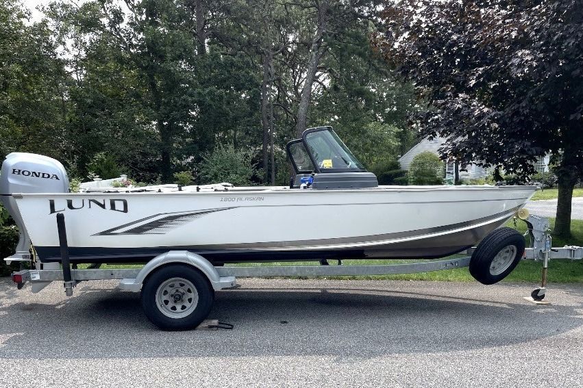 A white Lund boat, model Alask 2021, is on a trailer. It features a Honda outboard motor.