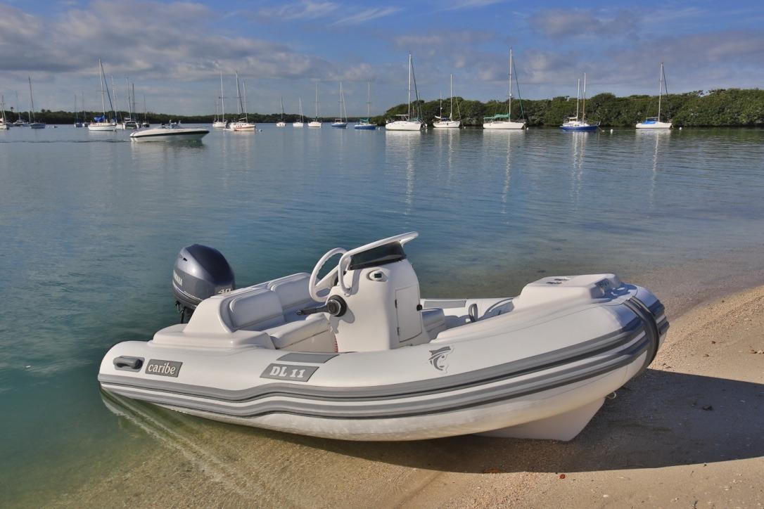 Inflatable Caribe DL11 boat on sandy shore, 2025 model, with sailboats in the background.