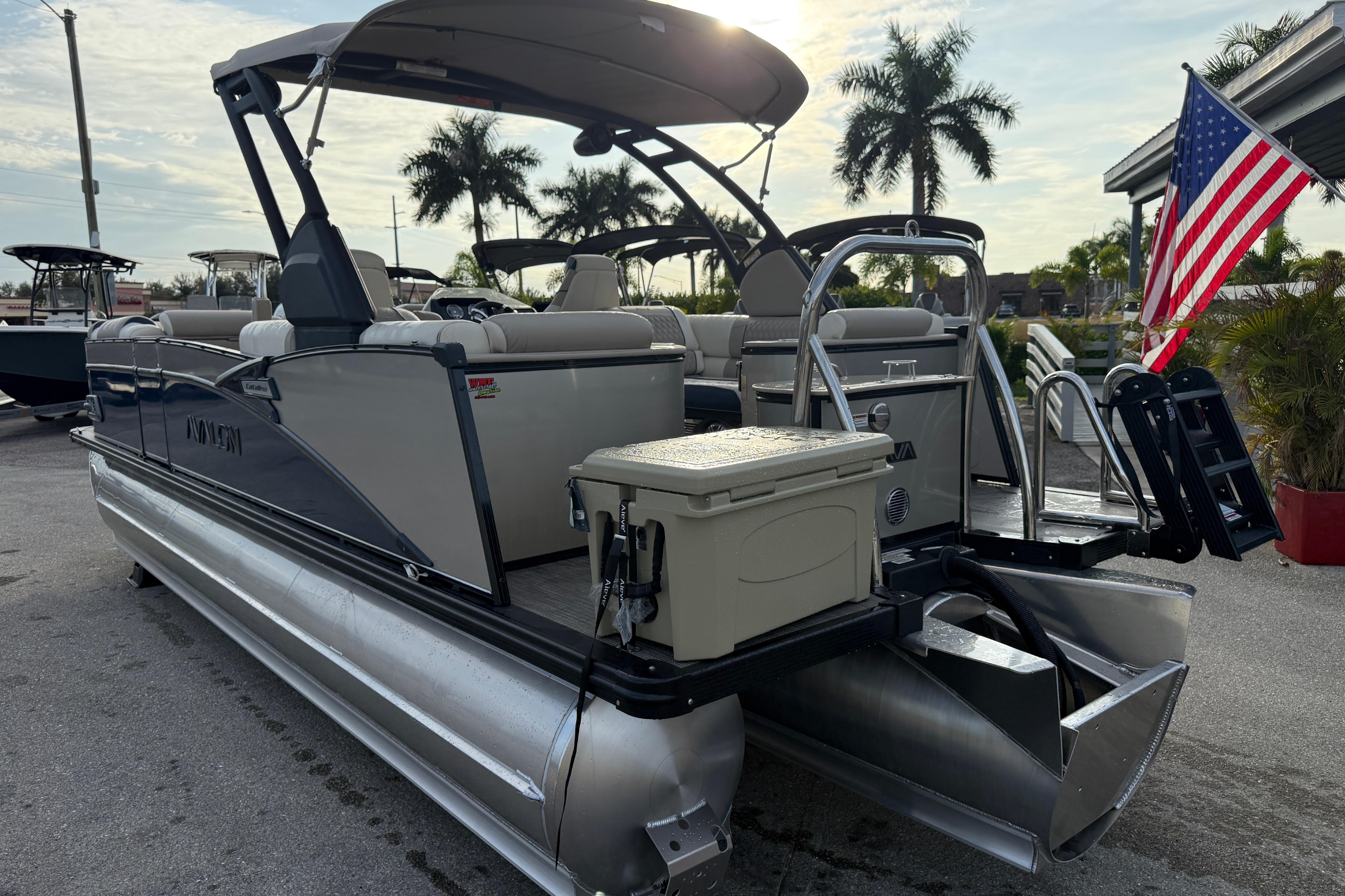 2026 Avalon 2385 Catalina Quad Lounger Tri Toon boat with American flag, parked outdoors.