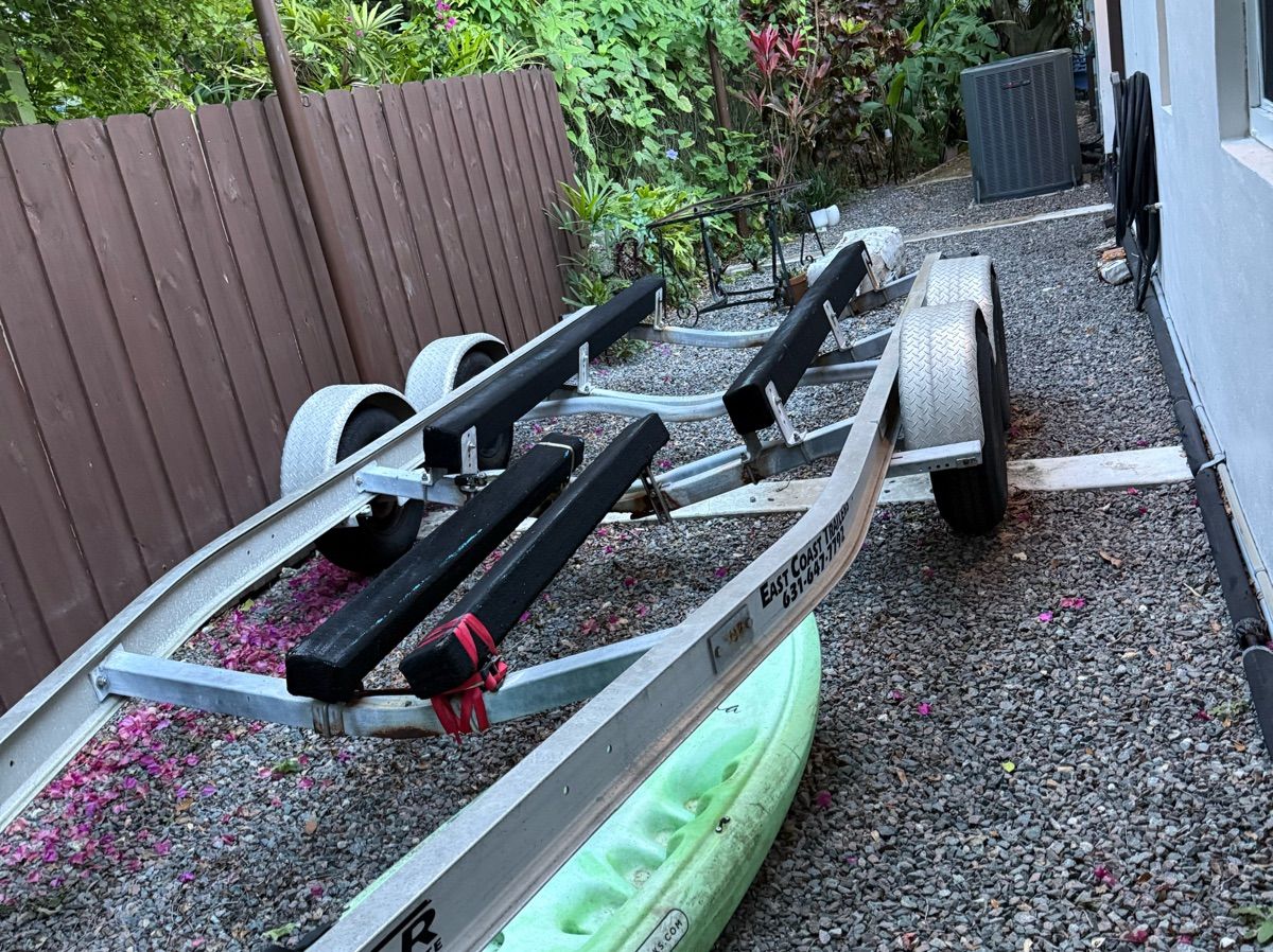 A boat trailer is parked on a gravel path next to a green kayak.