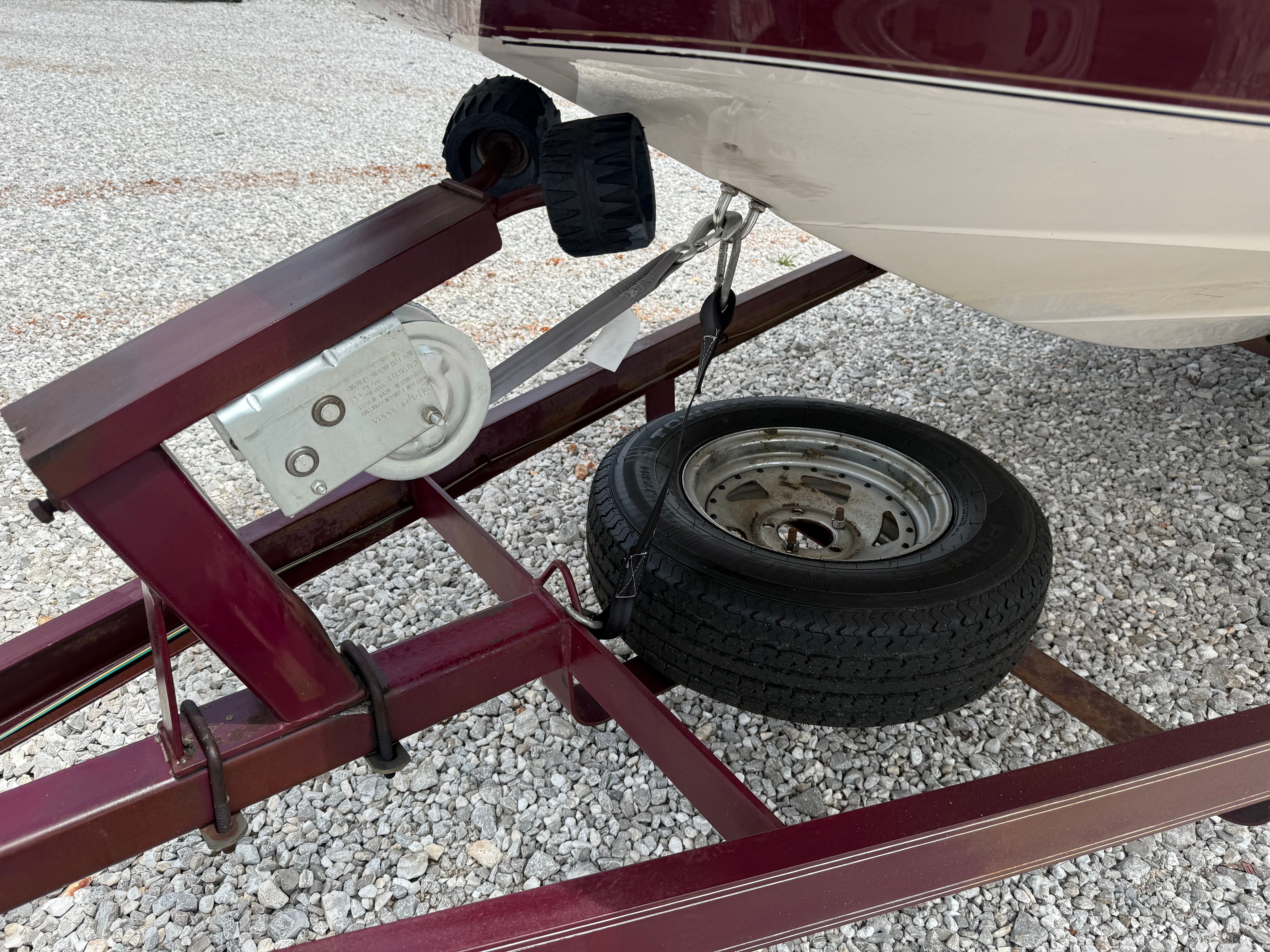 A boat trailer with a spare tire and winch on a gravel lot.