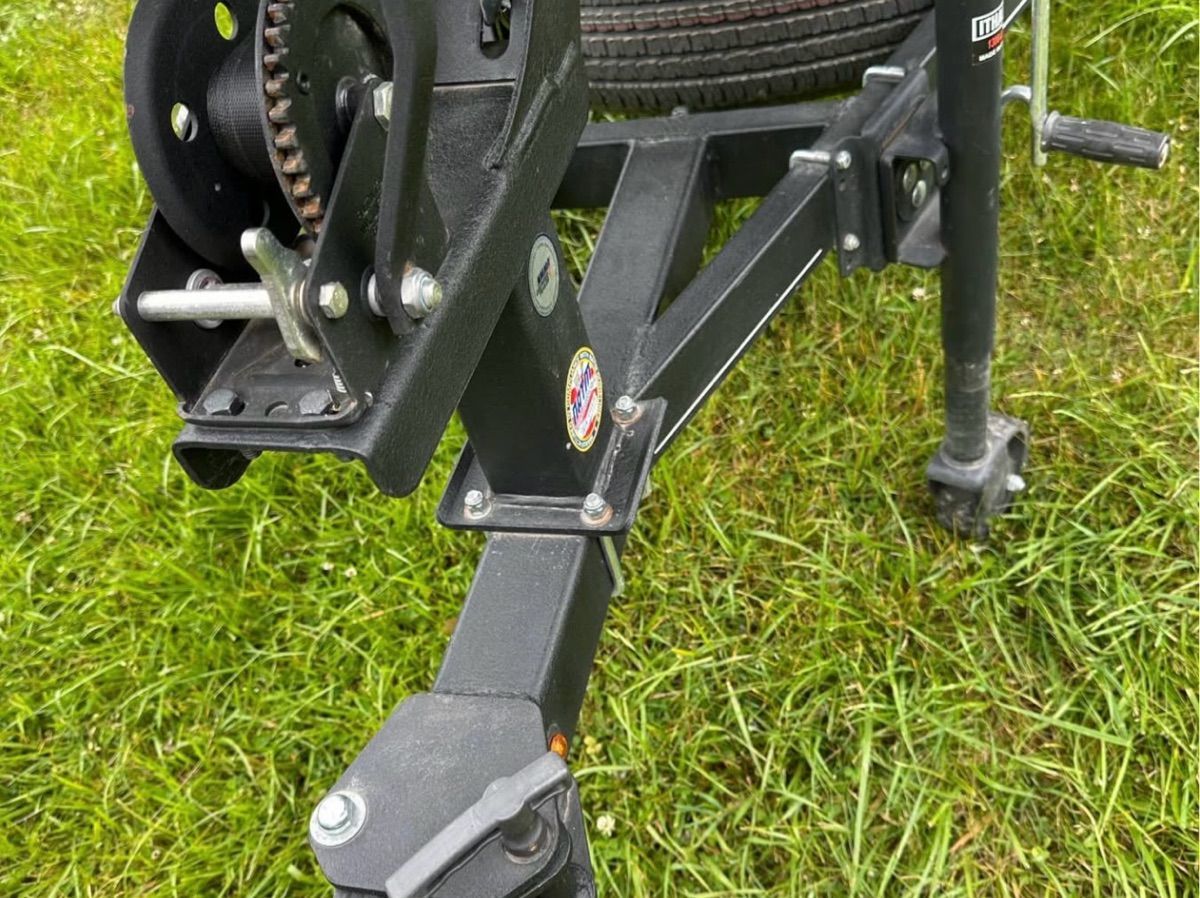 Close-up of a boat trailer with black metal frame and crank, standing on grass.