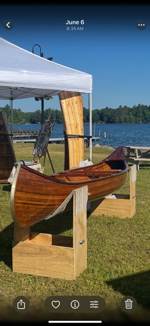 A handcrafted cedar strip canoe is displayed on a lawn, with a lake in the background.