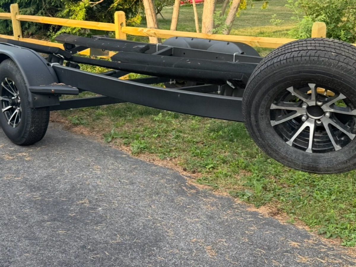 A black boat trailer is parked on a paved area next to a wooden fence.