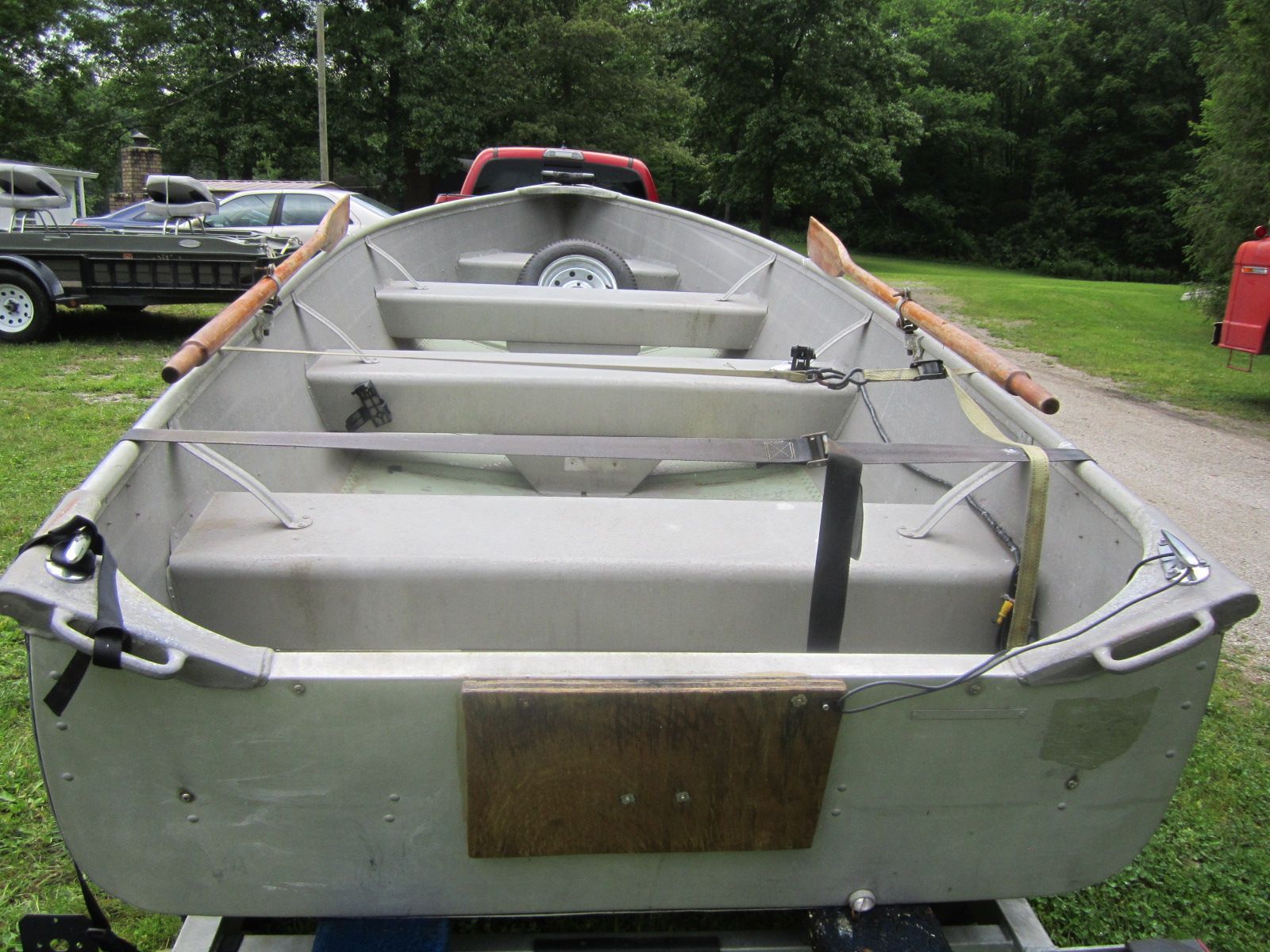 A gray rowboat on a trailer, surrounded by trees and vehicles.