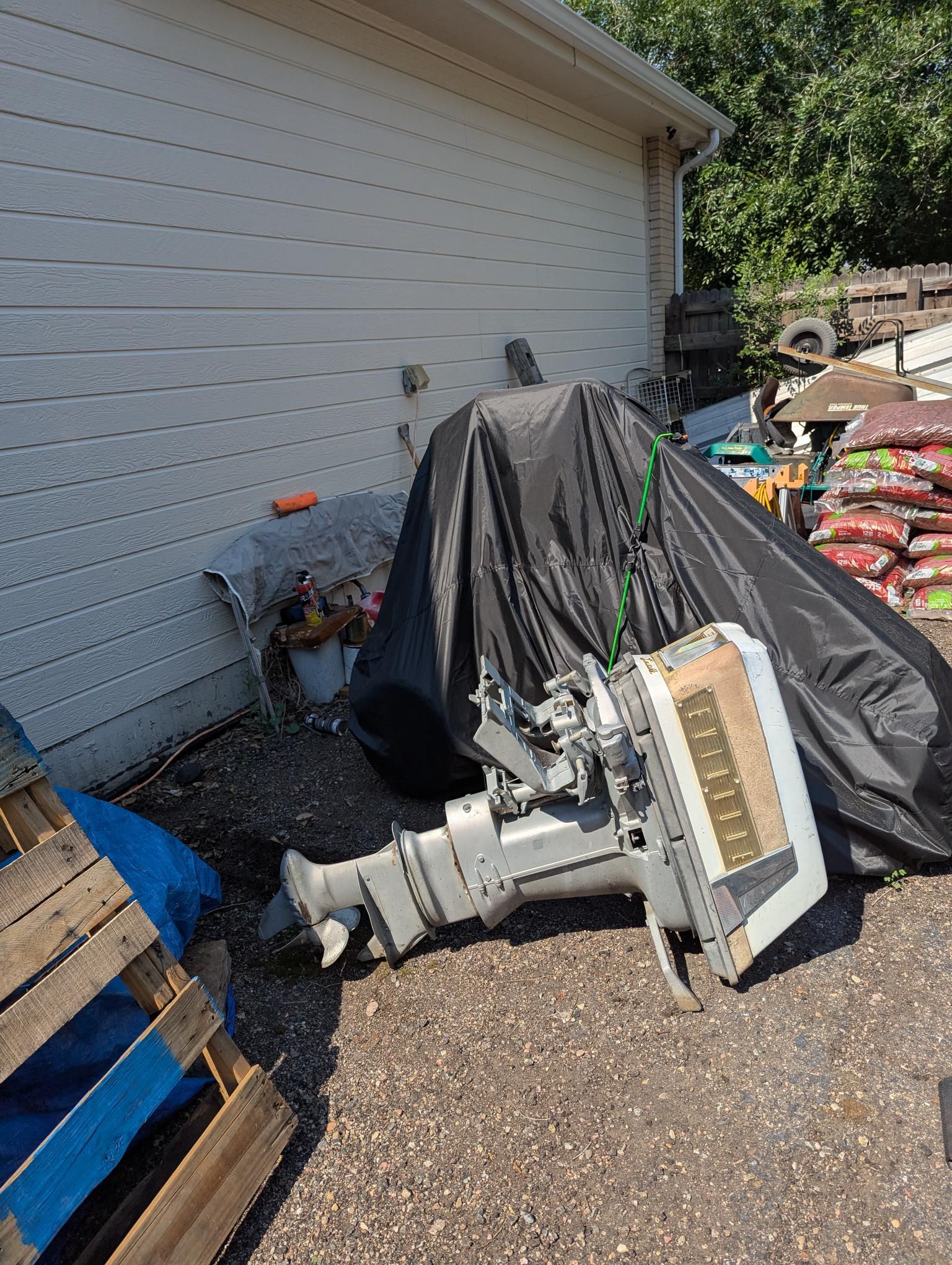 A covered boat next to a building, with an outboard motor on the ground.