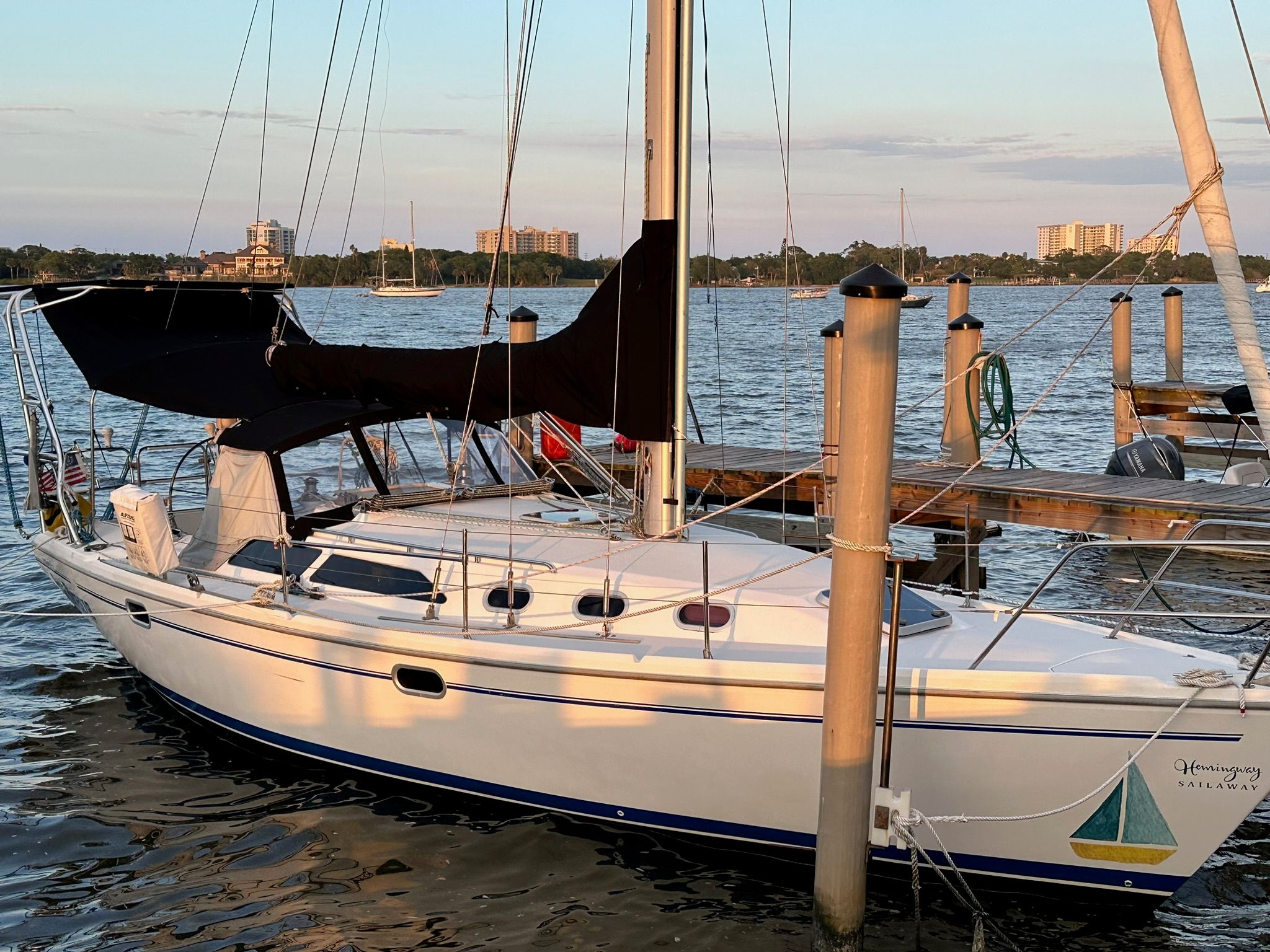 A white sailboat docked at a pier, surrounded by calm water and city buildings in the background.