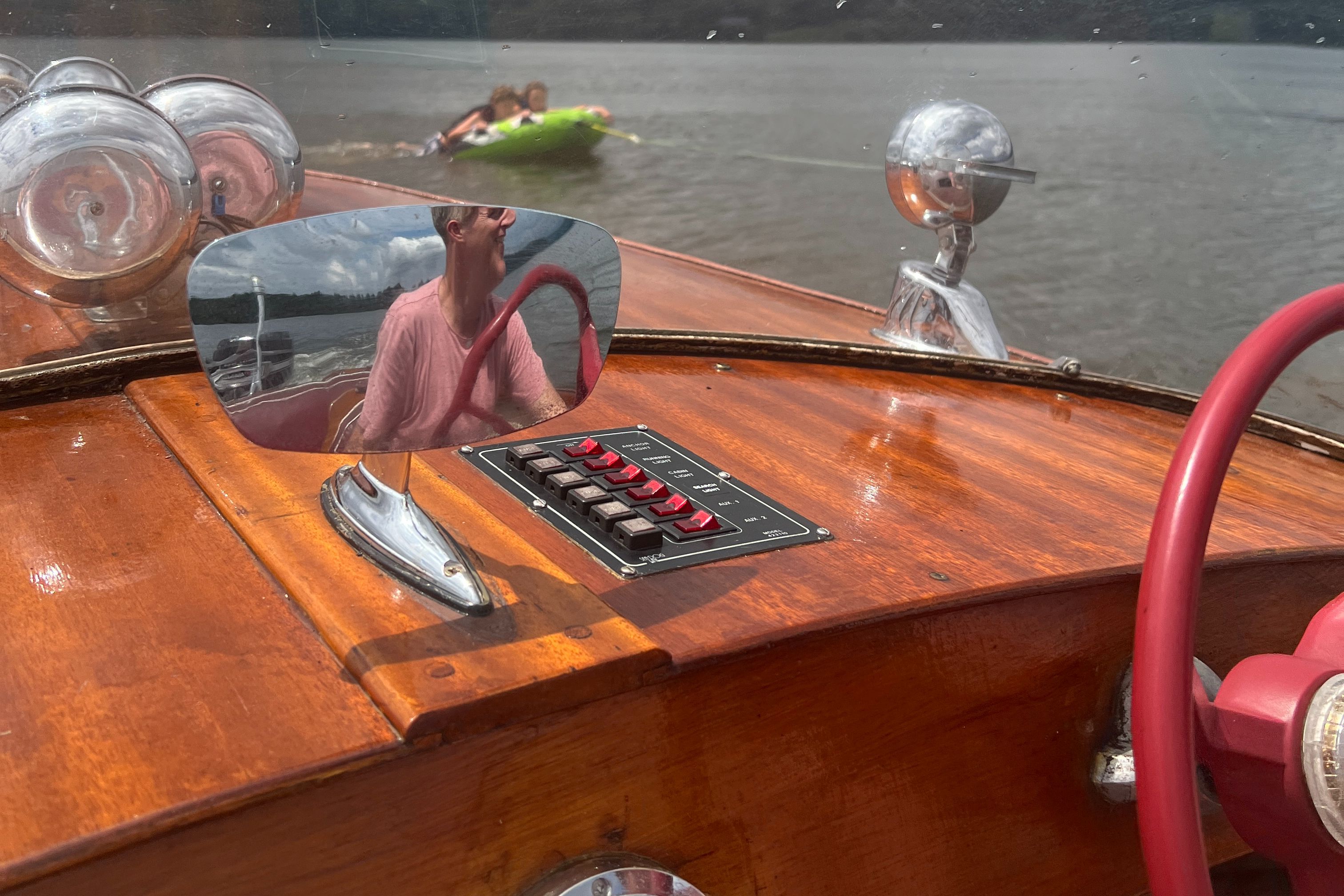 A classic wooden boat with a shiny dashboard and mirror. A tube is being towed in the background.