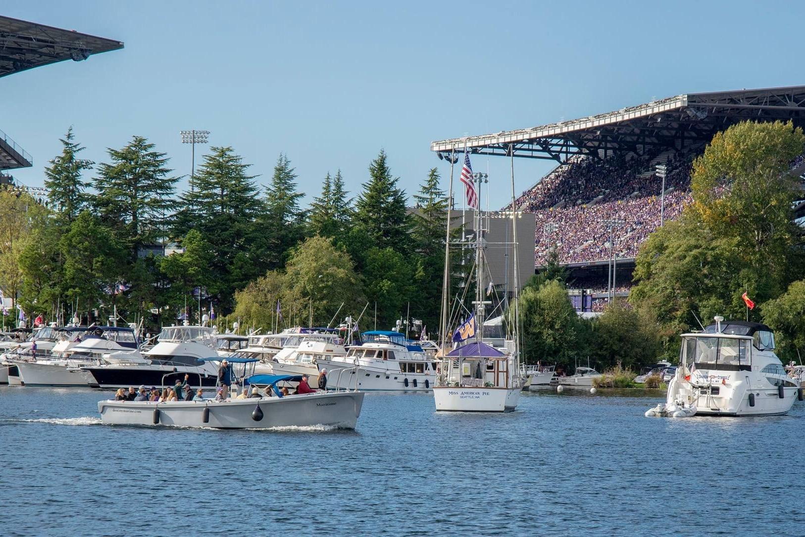 Boats on a lake near a crowded stadium, surrounded by lush trees under a clear blue sky.