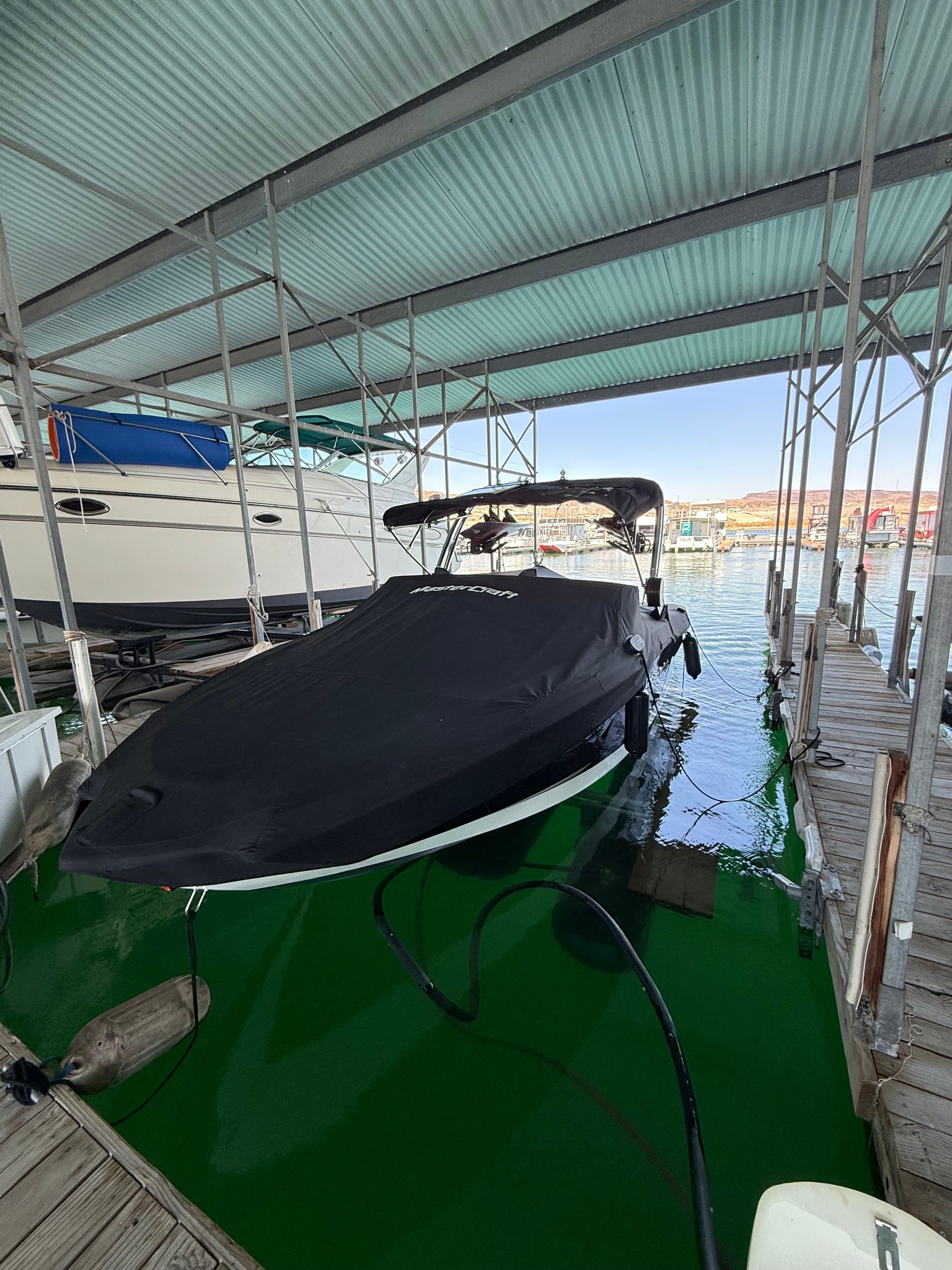 A MasterCraft NXT23 boat is docked under a covered area on green water.