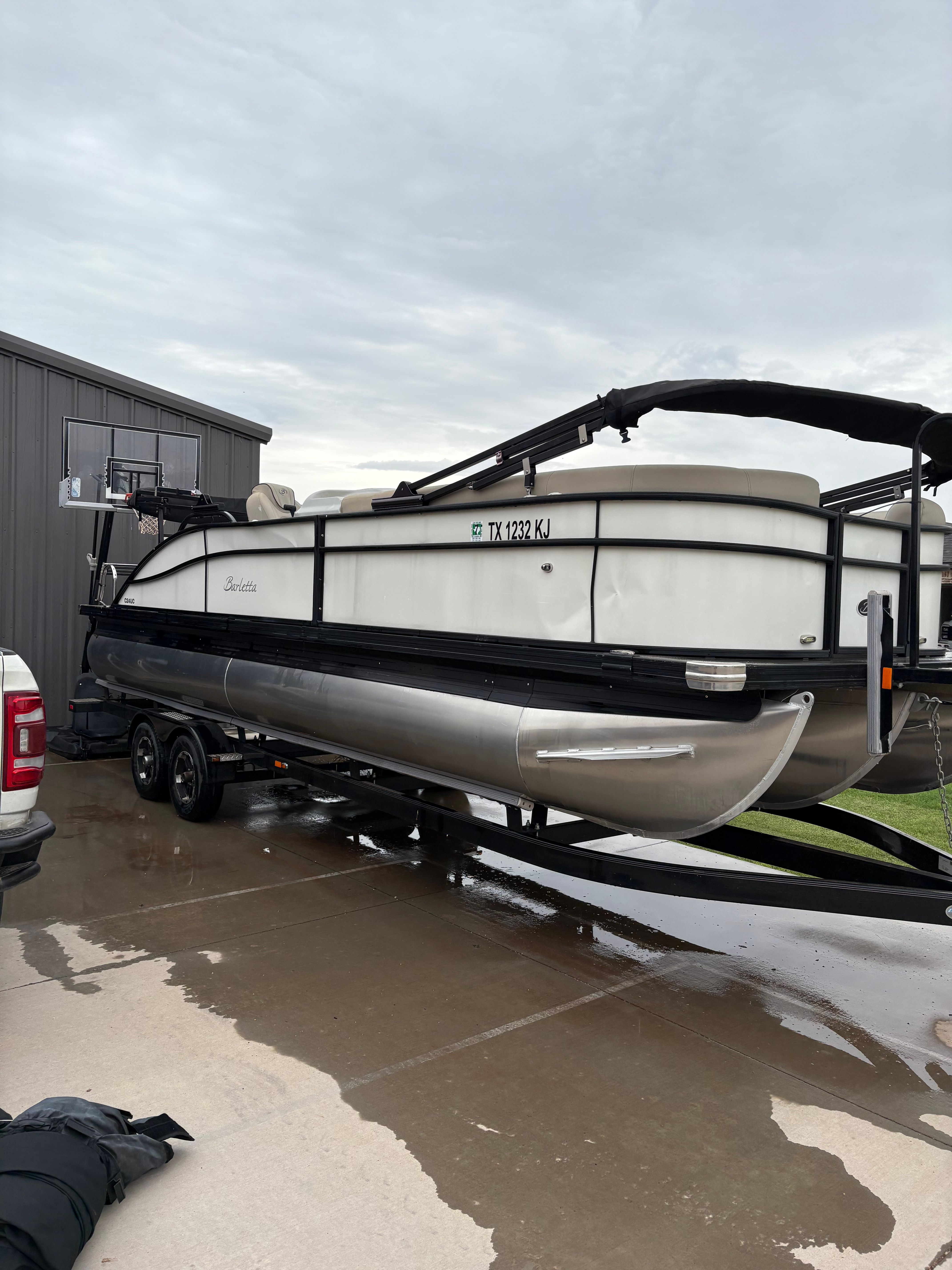 A Barletta pontoon boat on a trailer, parked on a wet driveway next to a building.