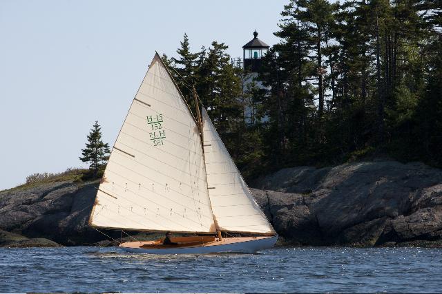 Sailboat Herreshoff Full-Keel Buzzards Bay 15 (2008) near rocky shore with lighthouse in background.