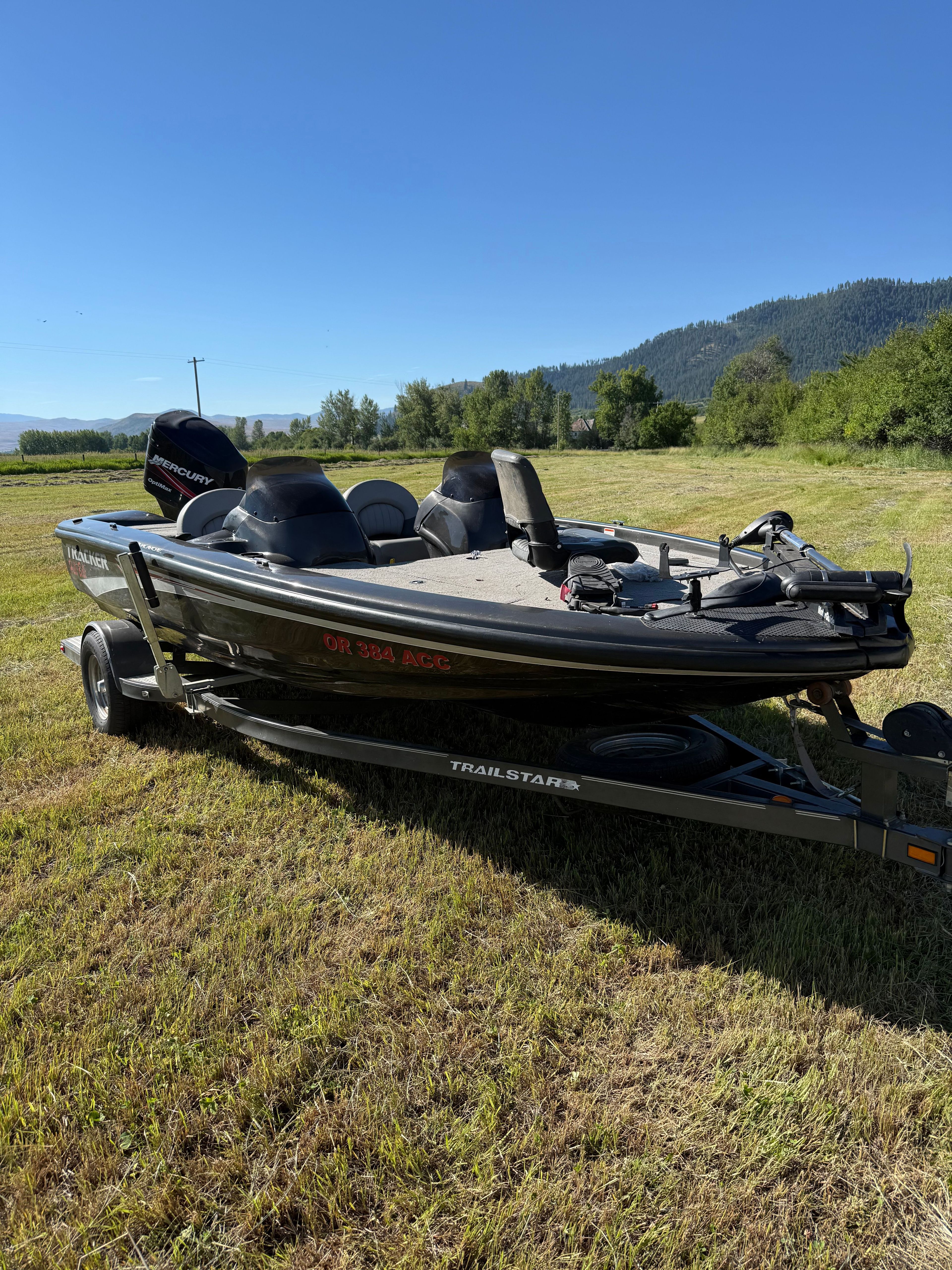 A black boat on a trailer sits on grass, surrounded by trees and mountains.
