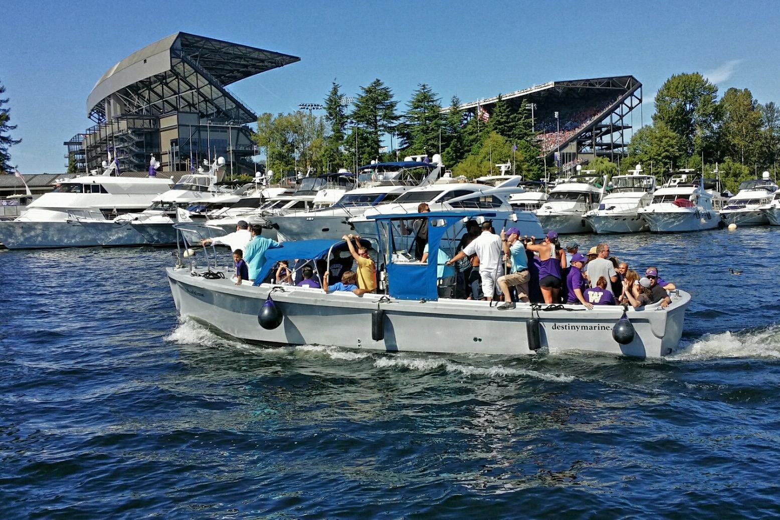 Boat with passengers on a lake near a stadium, surrounded by yachts, under a clear blue sky.