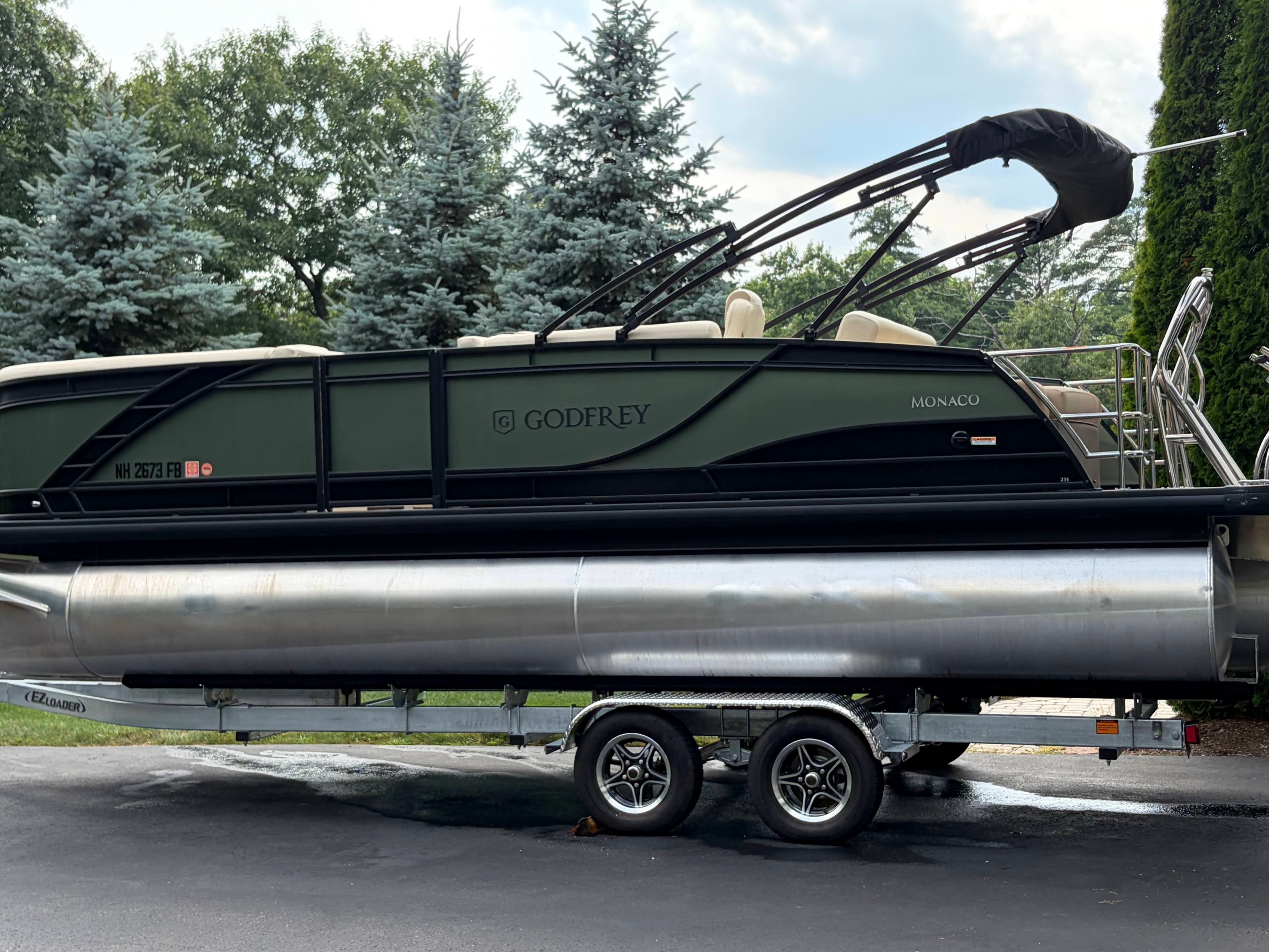 A 2025 Godfrey Monaco Cruise pontoon boat on a trailer, surrounded by trees.