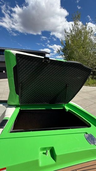 A green boat with an open storage hatch, surrounded by trees and blue sky.