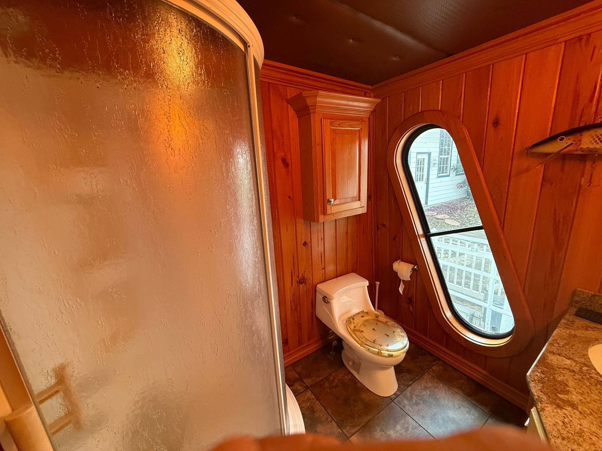 A bathroom with wood paneling, a shower, a toilet, and an oval window.