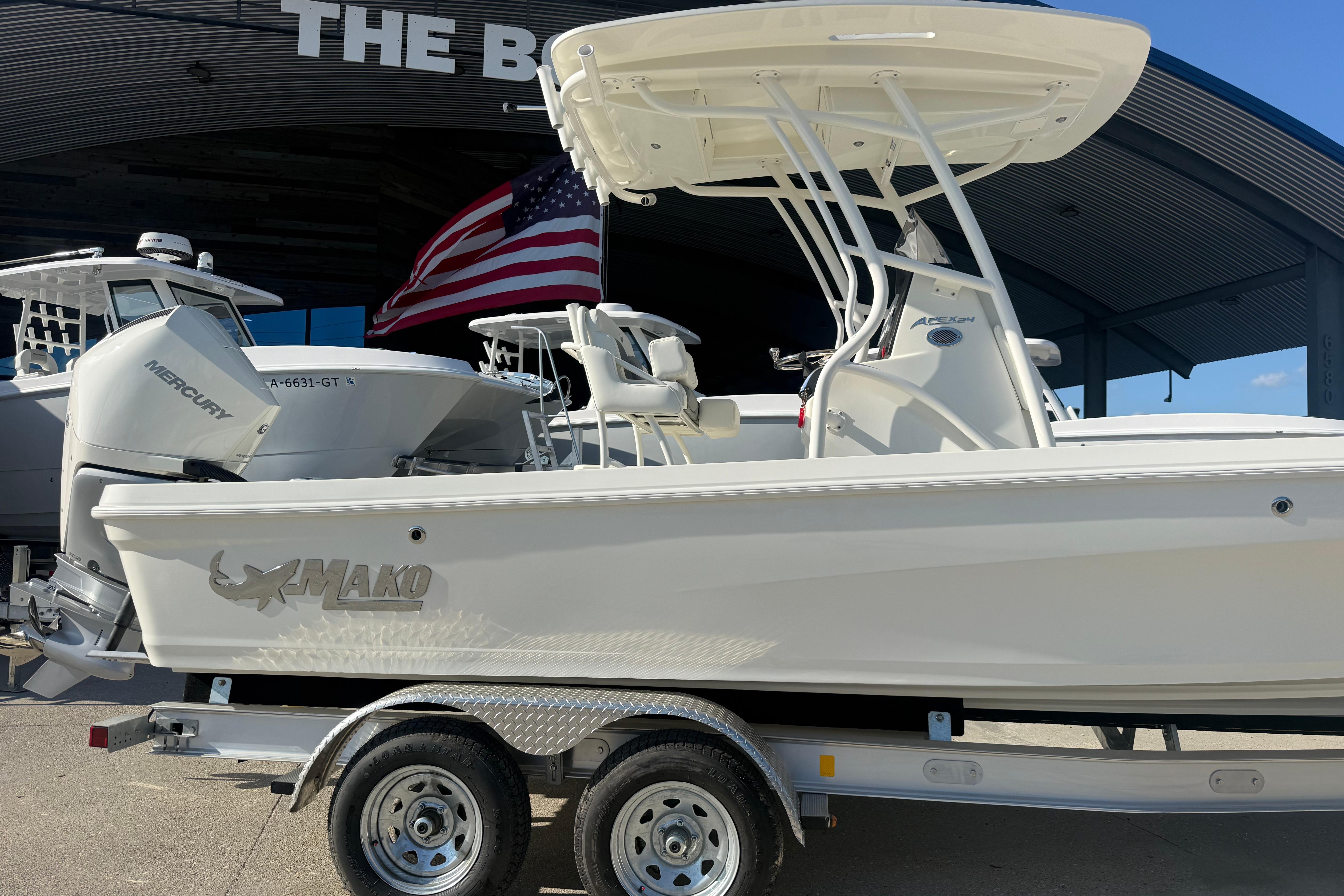 2026 Mako 24 APEX boat on trailer, American flag in background, under dealership awning.