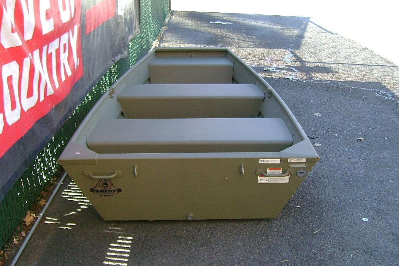 2026 Tracker Grizzly 10 Jon boat, olive green, parked on pavement near a fence.