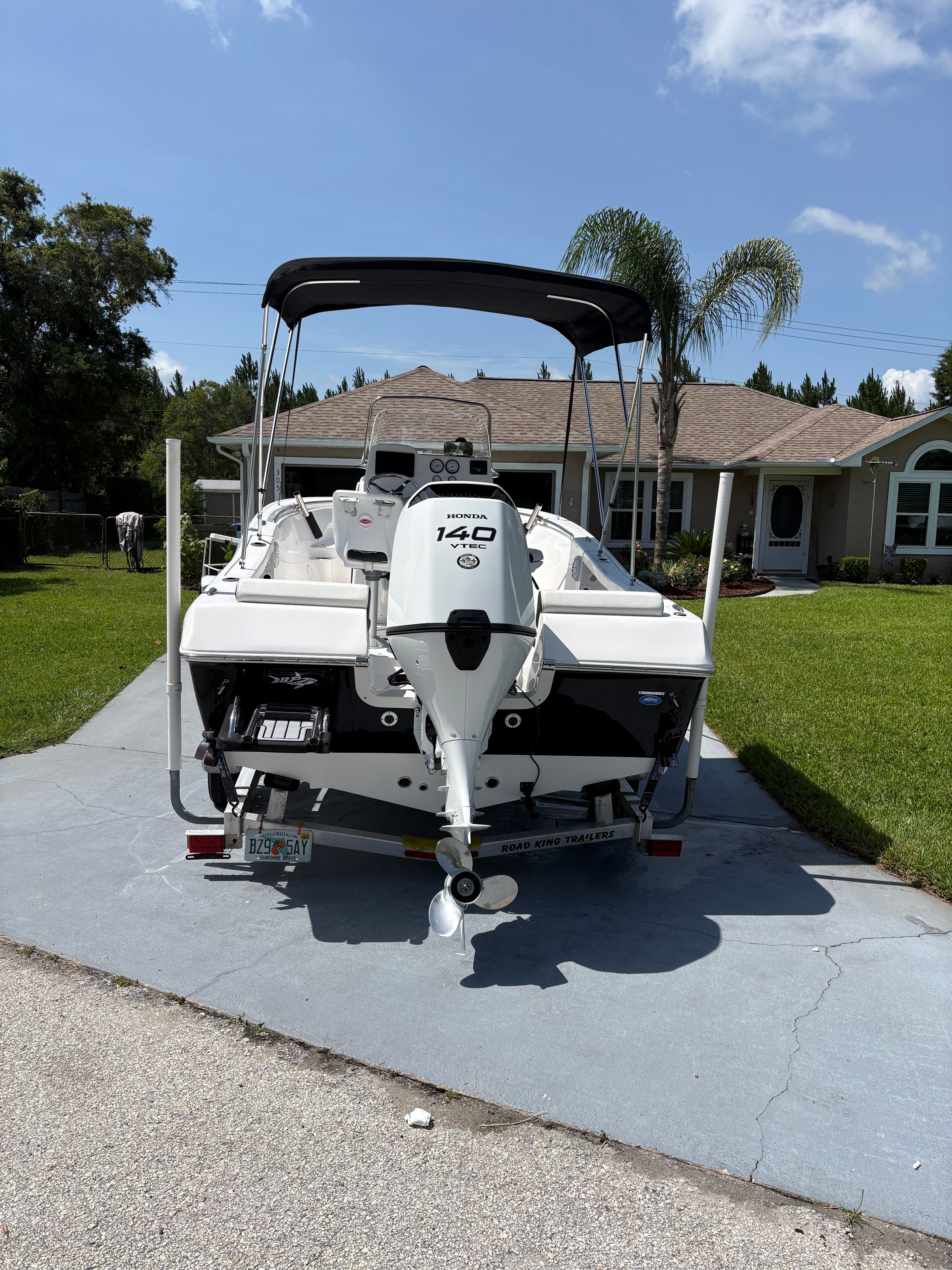 A white boat with an outboard motor sits on a driveway in front of a house.