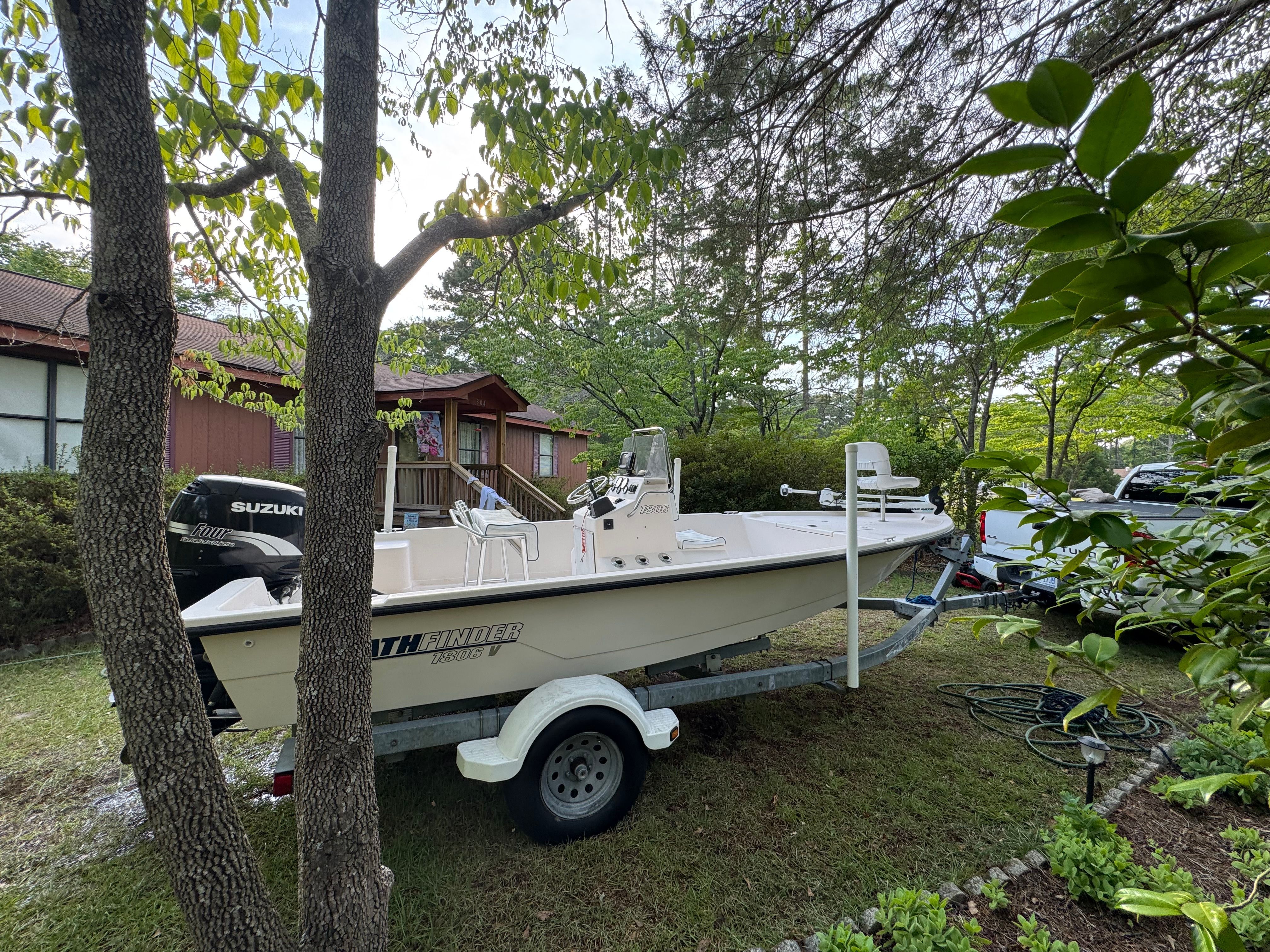 A white boat on a trailer is parked in a yard, surrounded by trees and a house.