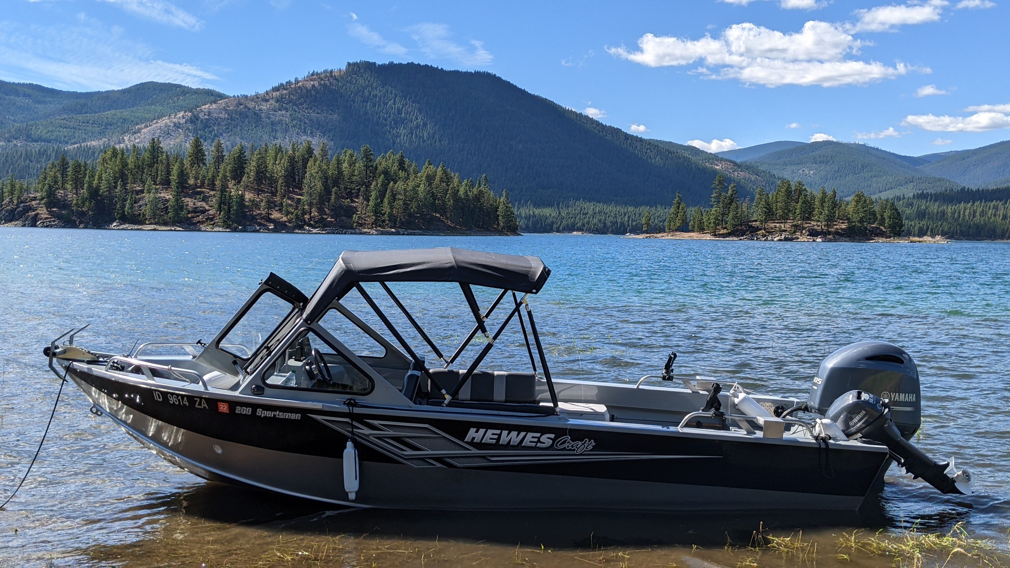 A 2020 Hewescraft 200 Sportsman boat is on a lake shore with forested hills in the background.
