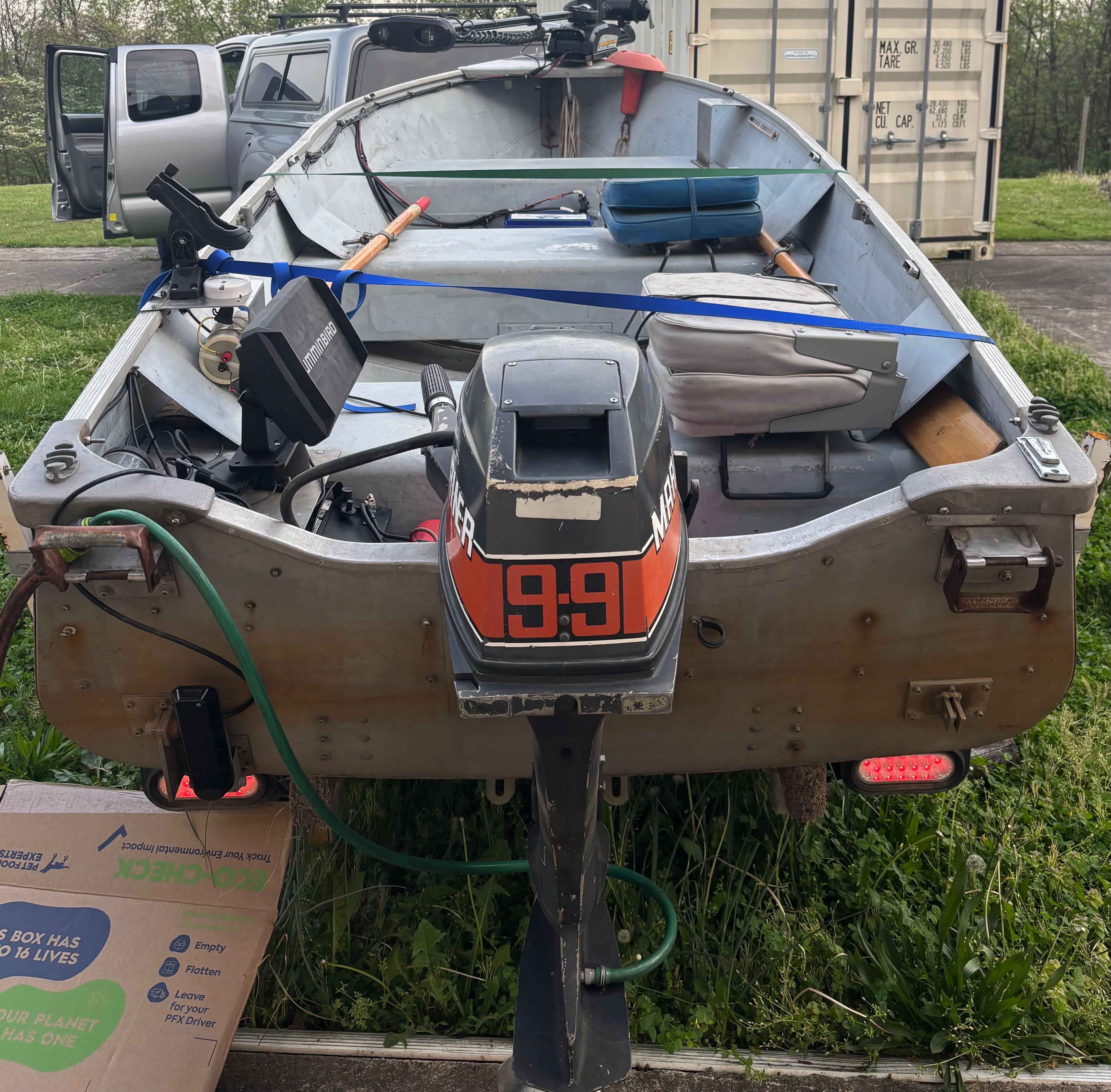 A 14-foot boat with an outboard motor, surrounded by grass and a container in the background.