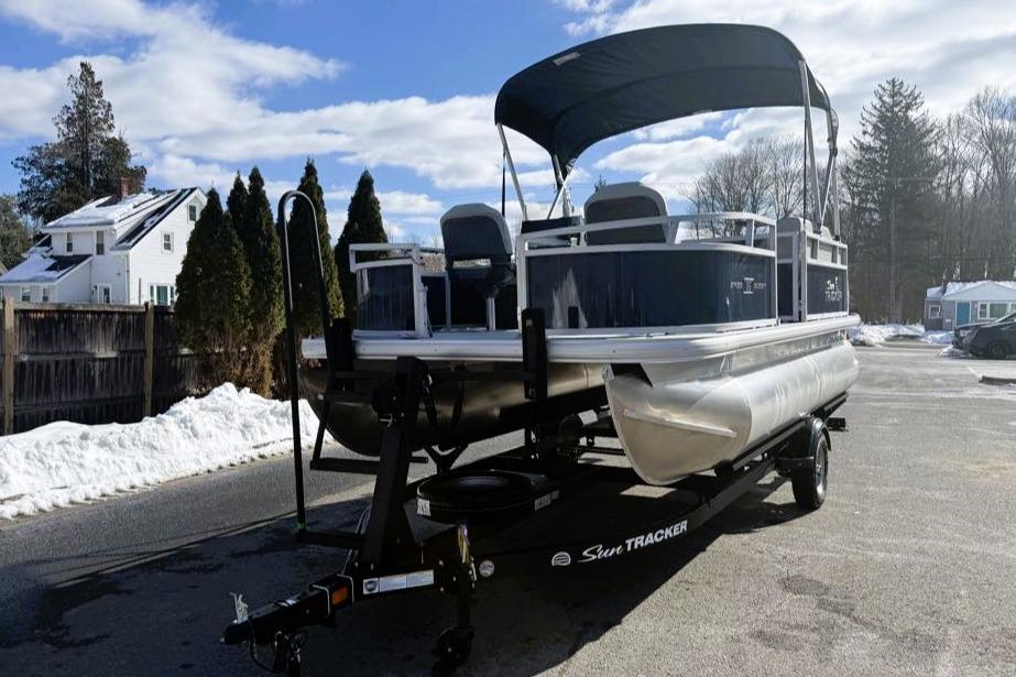 2026 Sun Tracker Bass Buggy 16 XL Select pontoon boat on trailer, parked in snowy driveway.