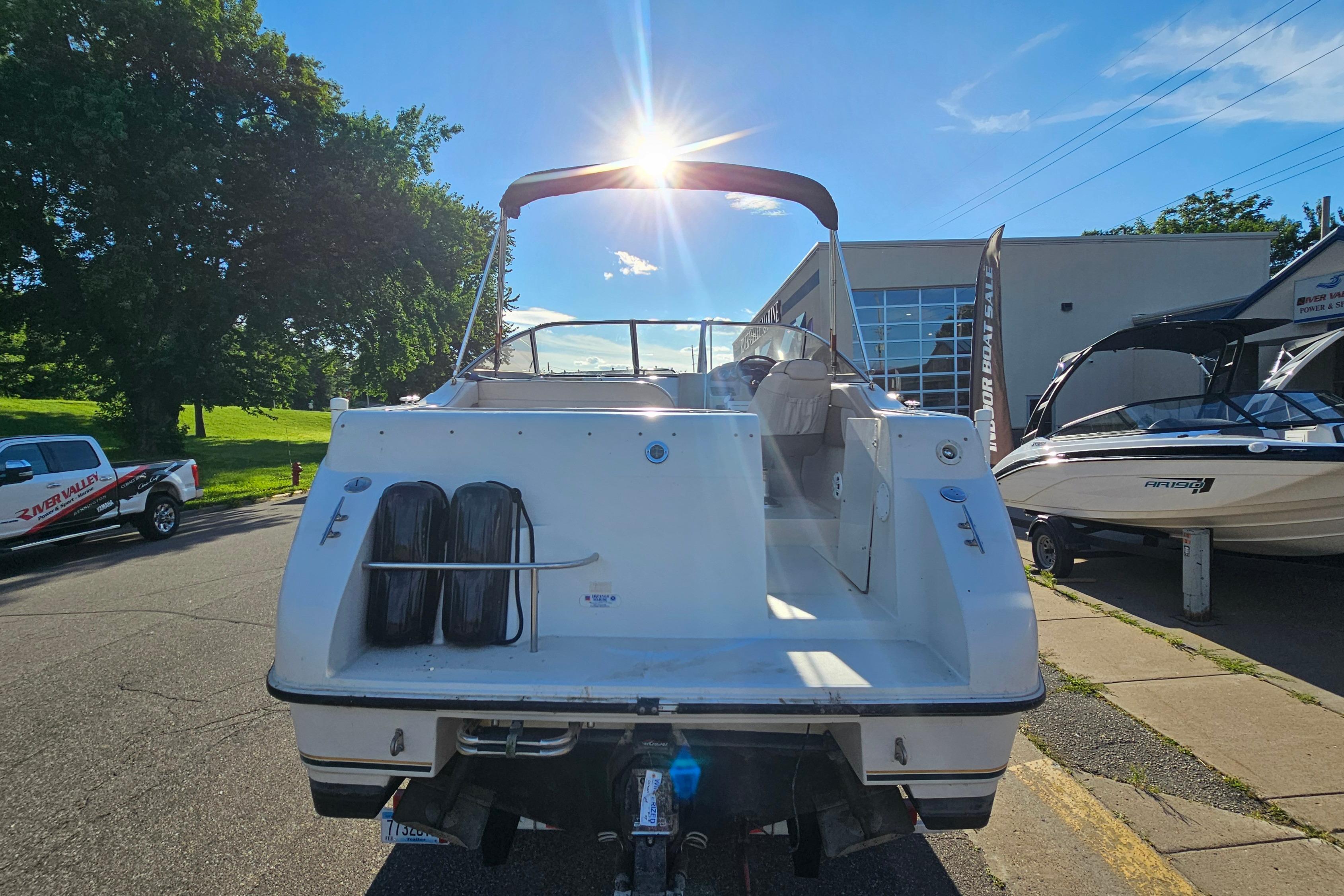 1999 Regal Commodore 258 boat parked outdoors, sun shining above, surrounded by trees and vehicles.