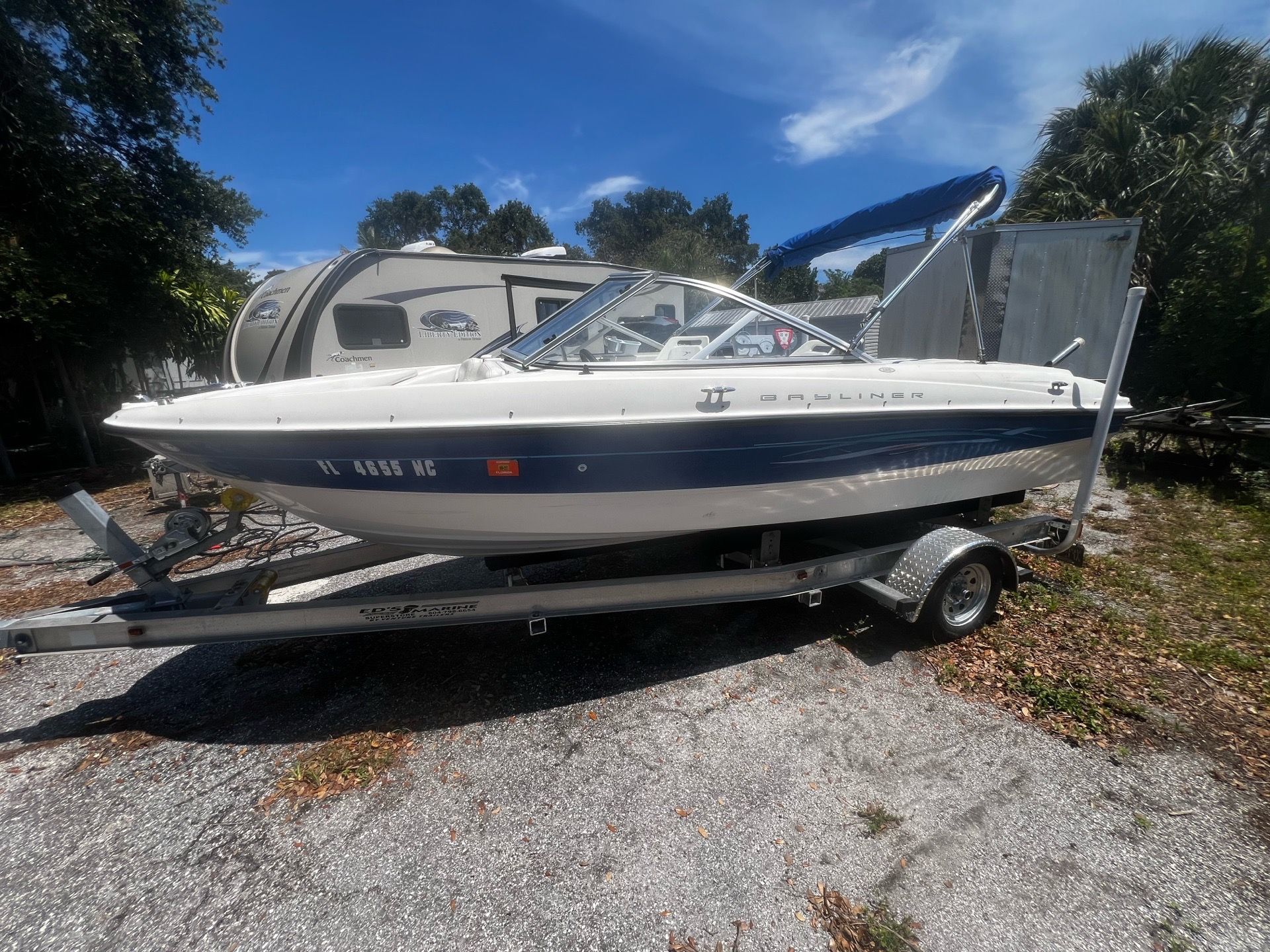 A 2006 Bayliner 185 boat on a trailer, surrounded by trees and a camper.