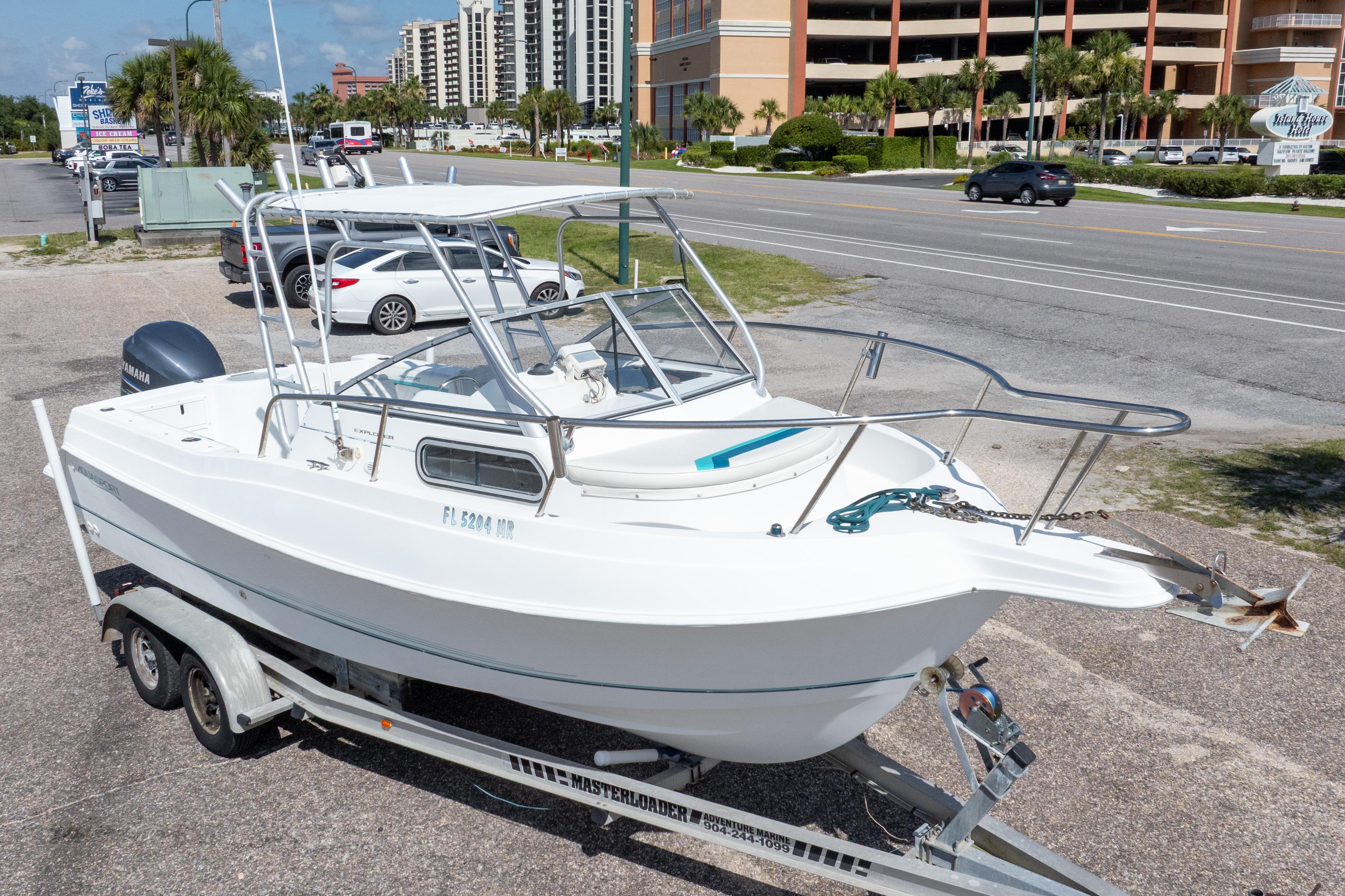 1999 Aquasport 225 Explorer boat on trailer, parked near a road with buildings in background.