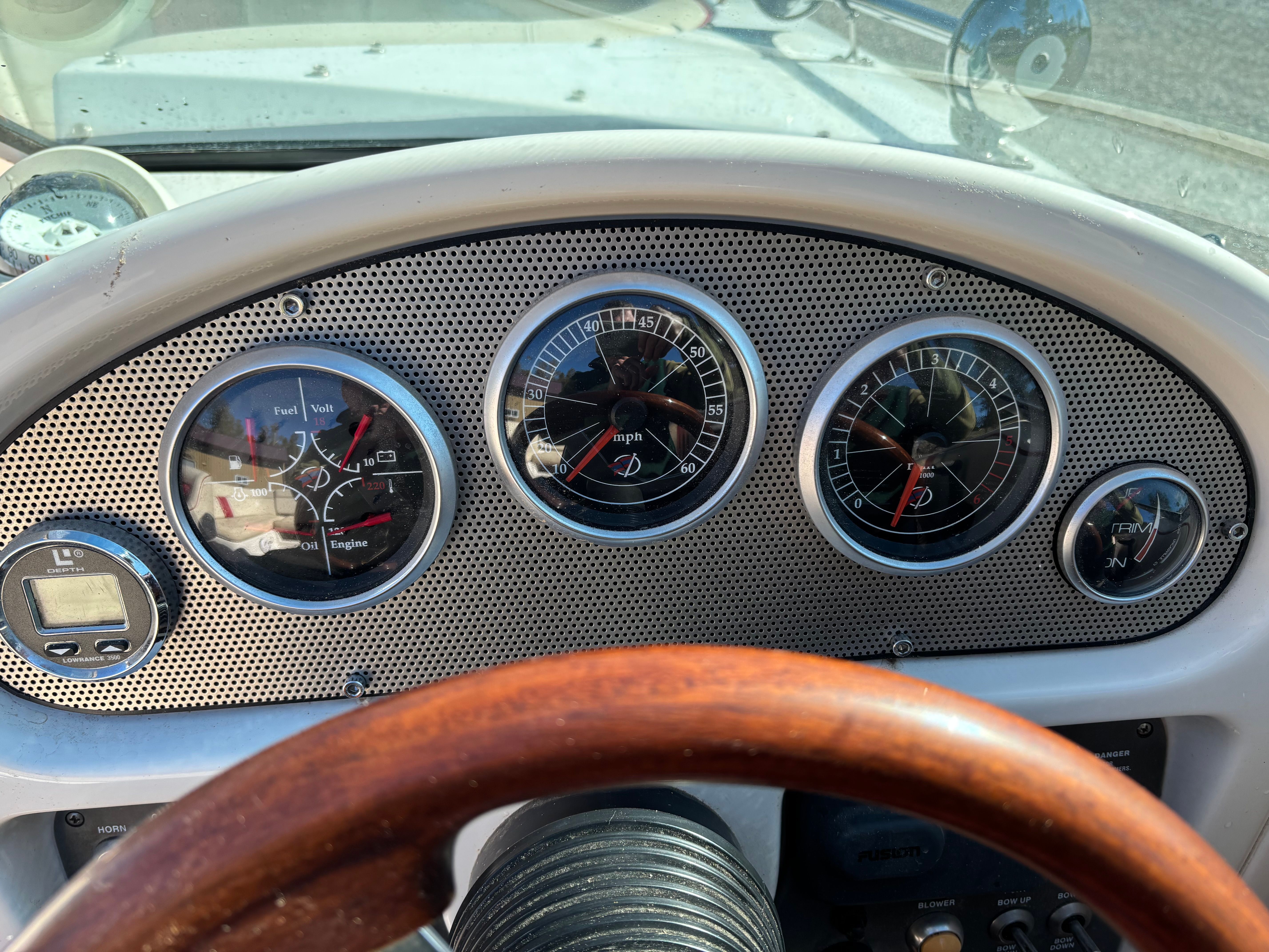 The image shows a boat dashboard with several gauges and a wooden steering wheel.