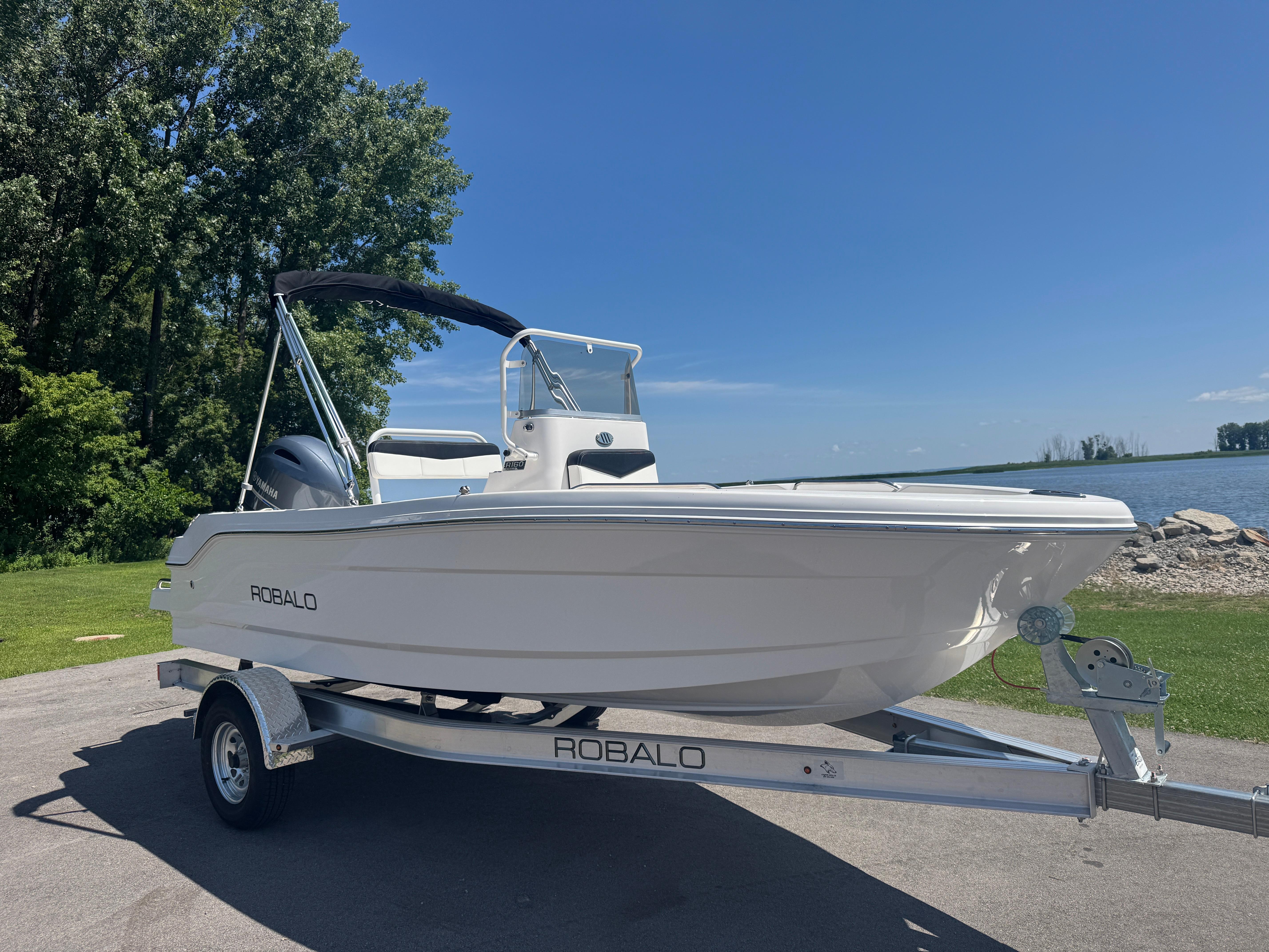 2025 Robalo R160 Center Console boat on trailer by a lake under clear blue sky.