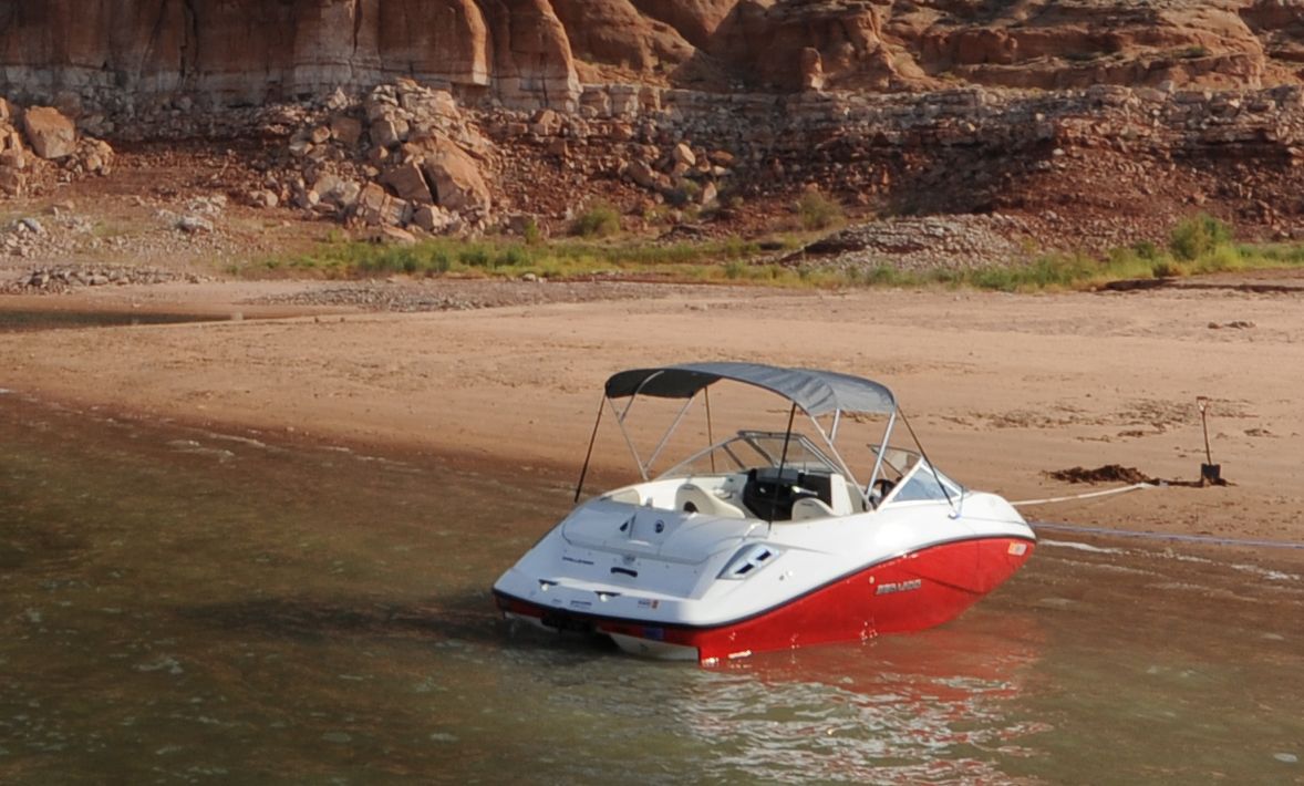 A white and red boat with a gray sunshade is docked at a sandy lakeshore.