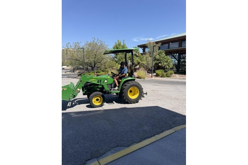 Person driving a green tractor on a sunny day, surrounded by trees and buildings.