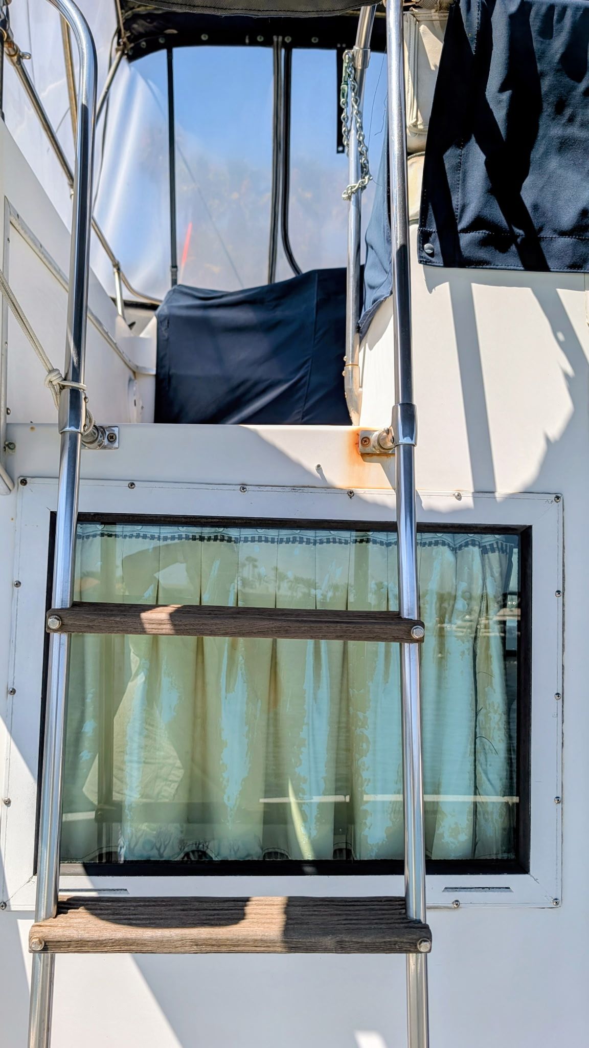A ladder leads to an elevated area on a boat. Below is a window with curtains.