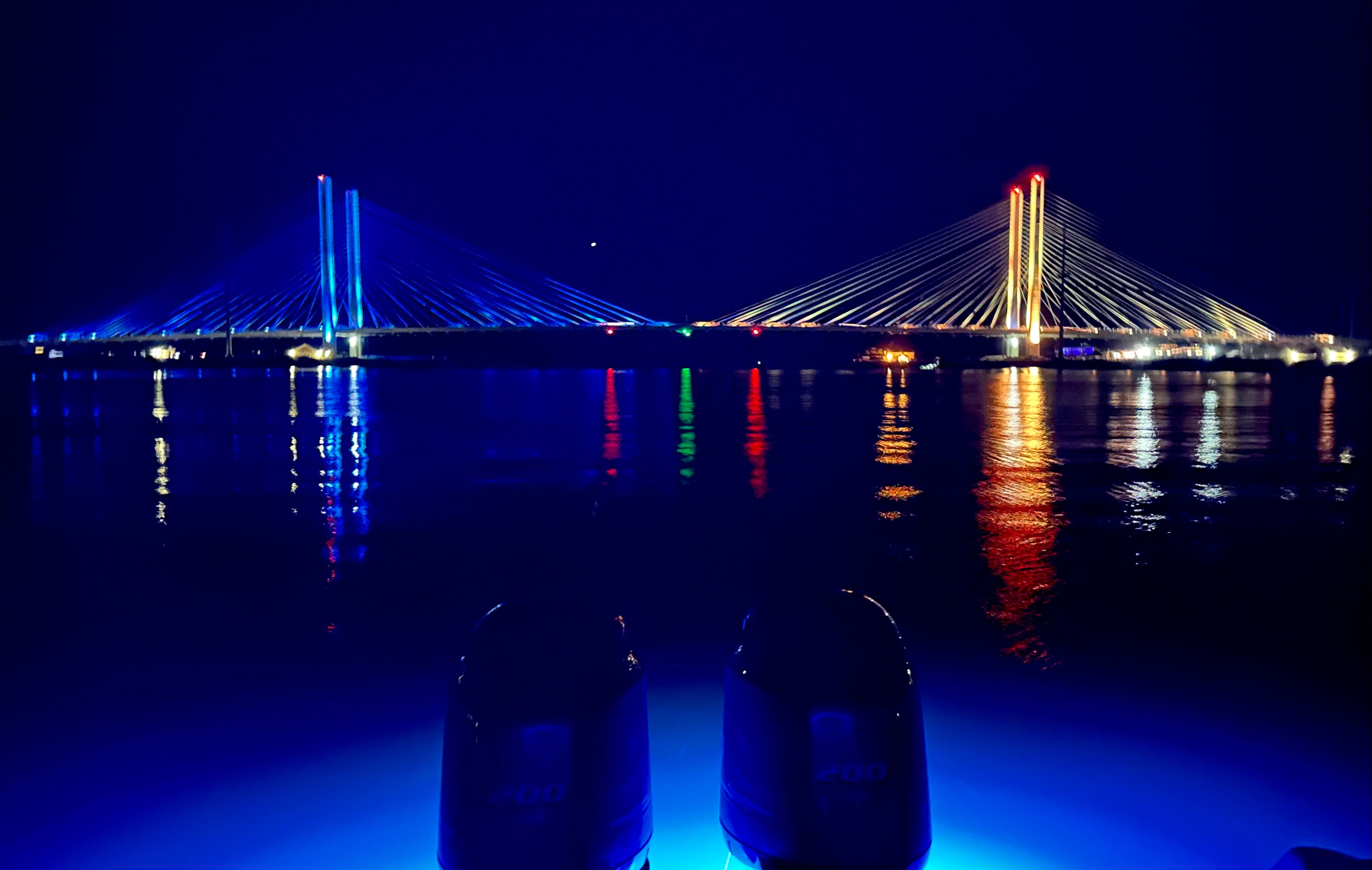 Night view of two illuminated bridges over calm water, with a boat in the foreground.