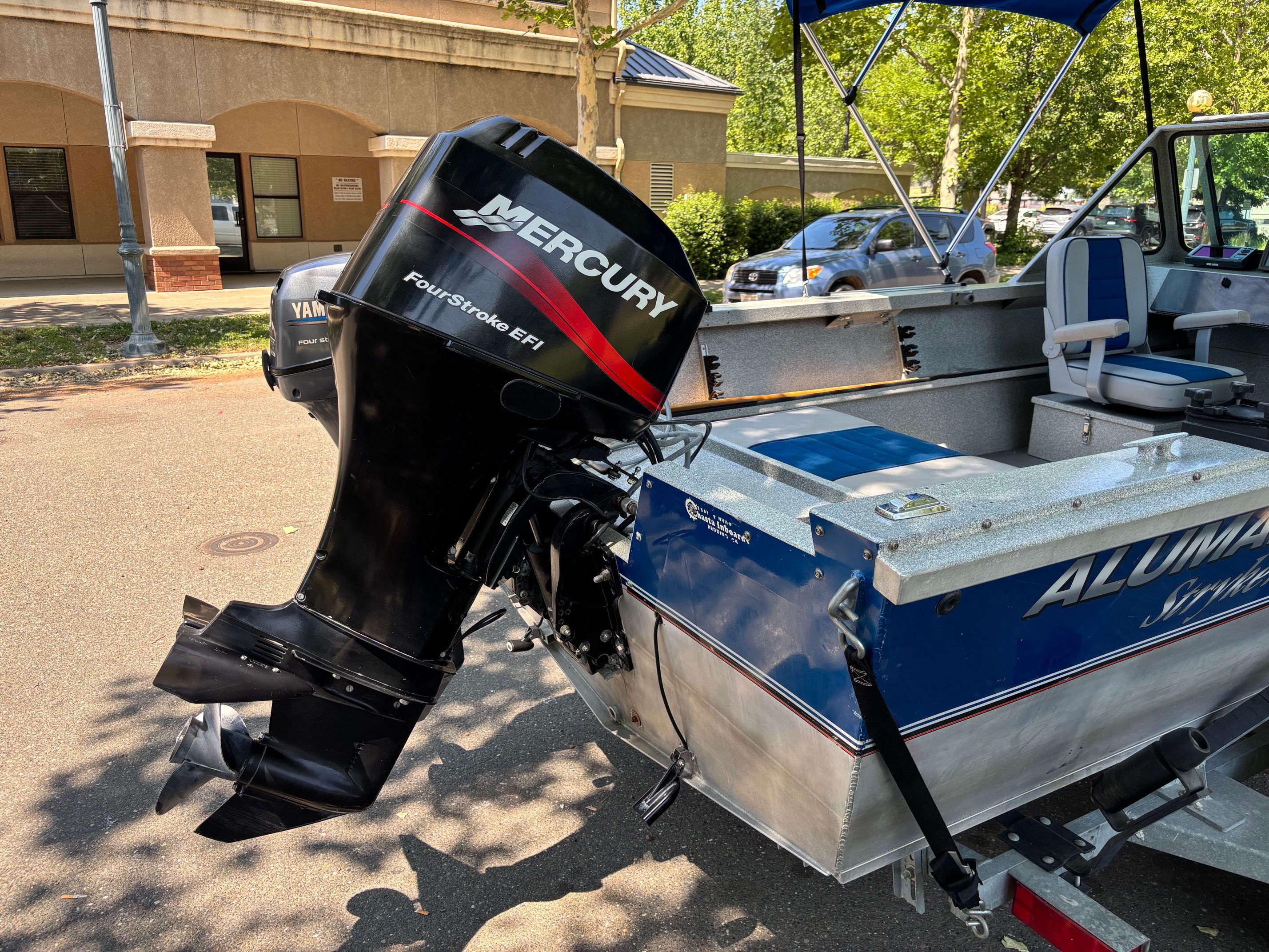 A blue boat with a Mercury outboard motor, parked on a trailer in an urban setting.