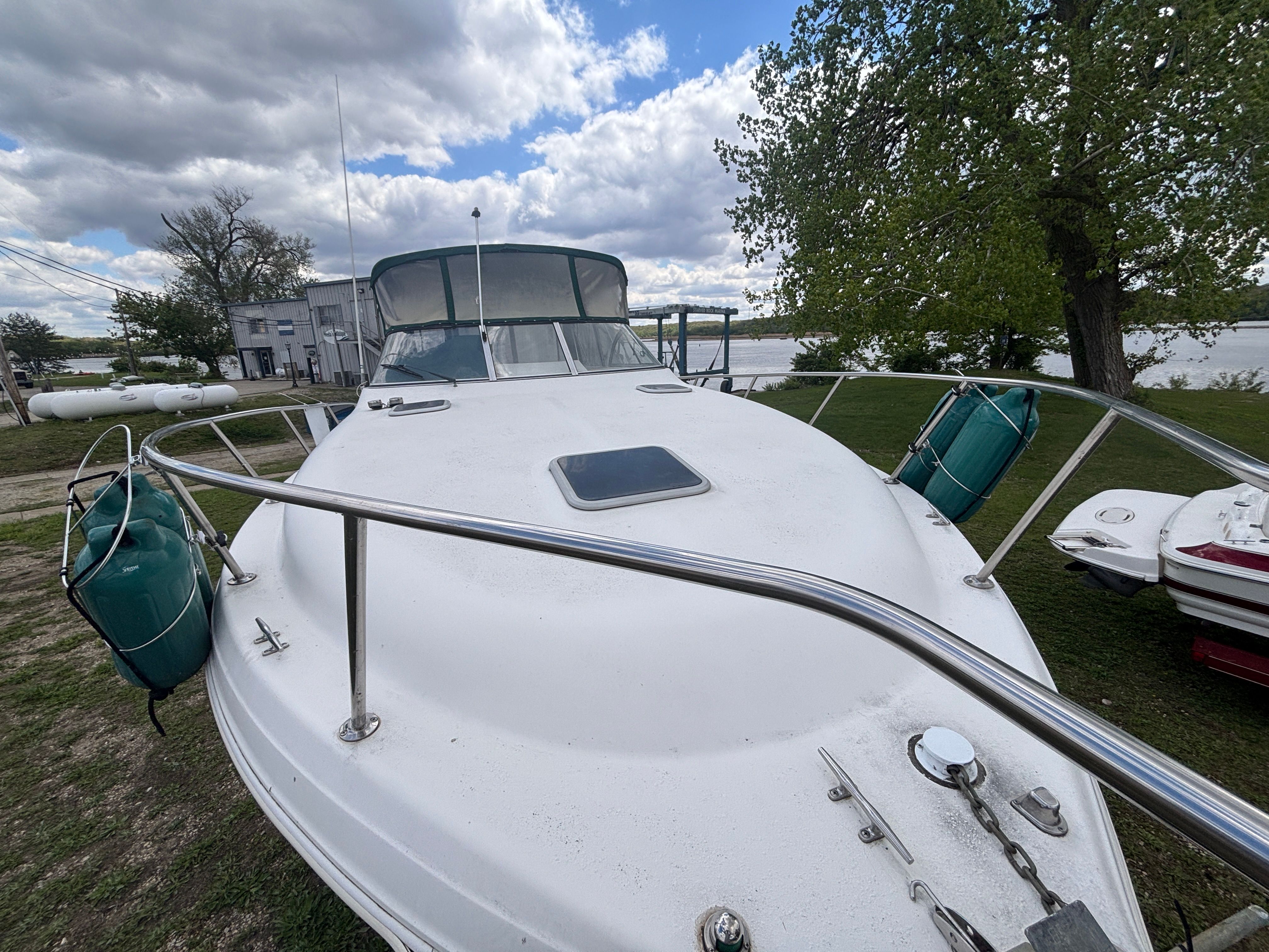 A white boat is on the grass, surrounded by trees and a building in the background.