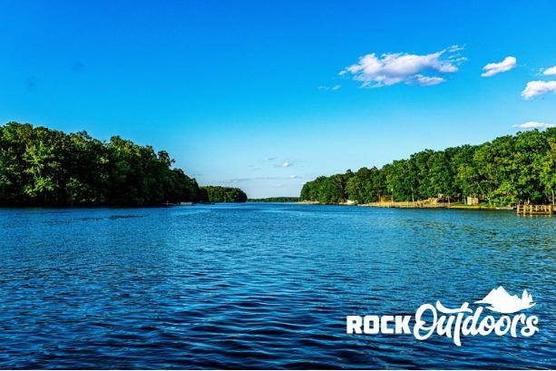 A serene lake view with lush green trees under a clear blue sky, Rock Outdoors logo visible.