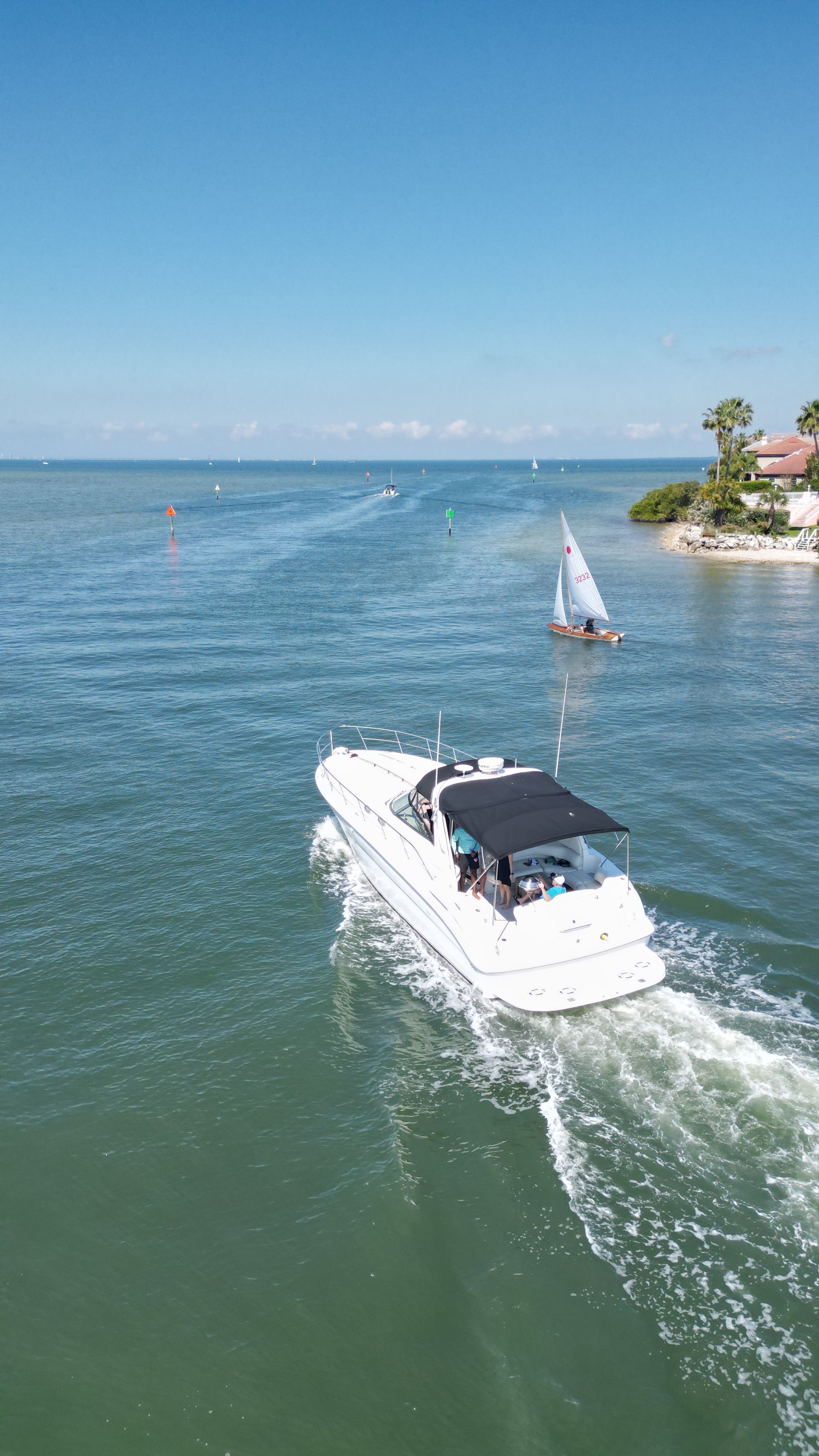 A white motorboat cruises on calm water, with a sailboat nearby and a coastline in the background.