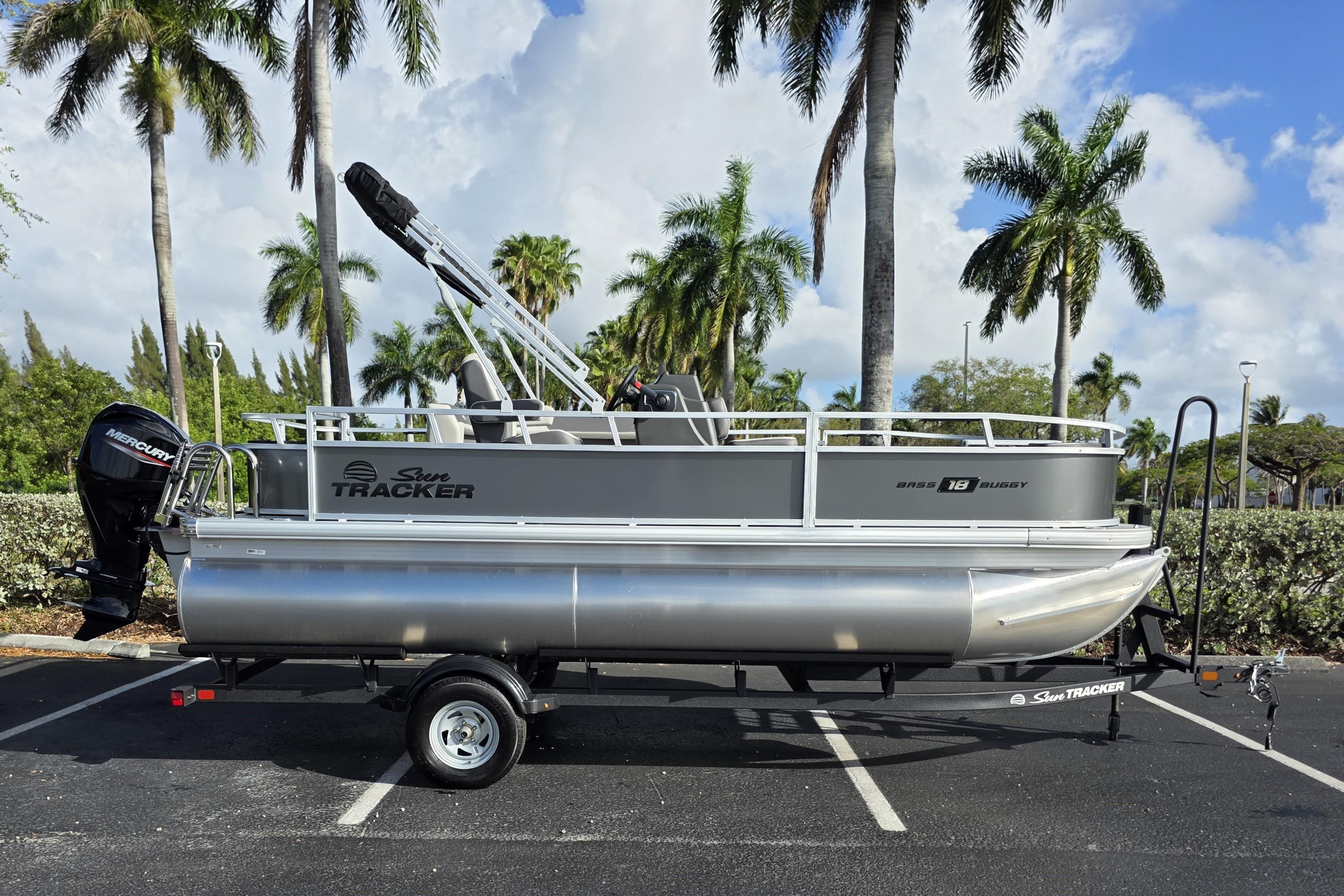 2026 Sun Tracker Bass Buggy 16 XL Select pontoon boat on trailer, parked under palm trees.