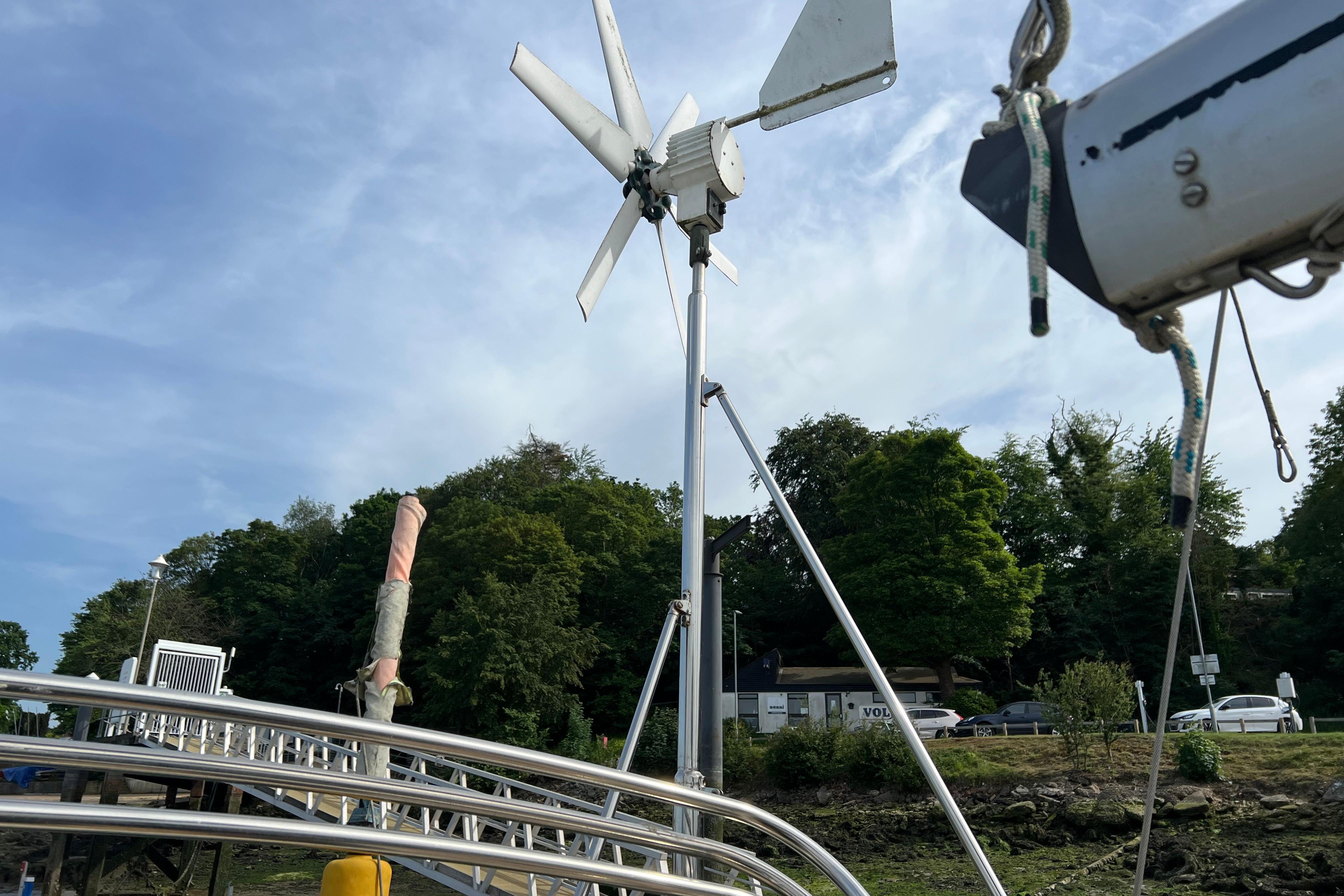 Wind turbine on a 1996 Bruce Roberts Cutter sailboat, docked near a lush shoreline.