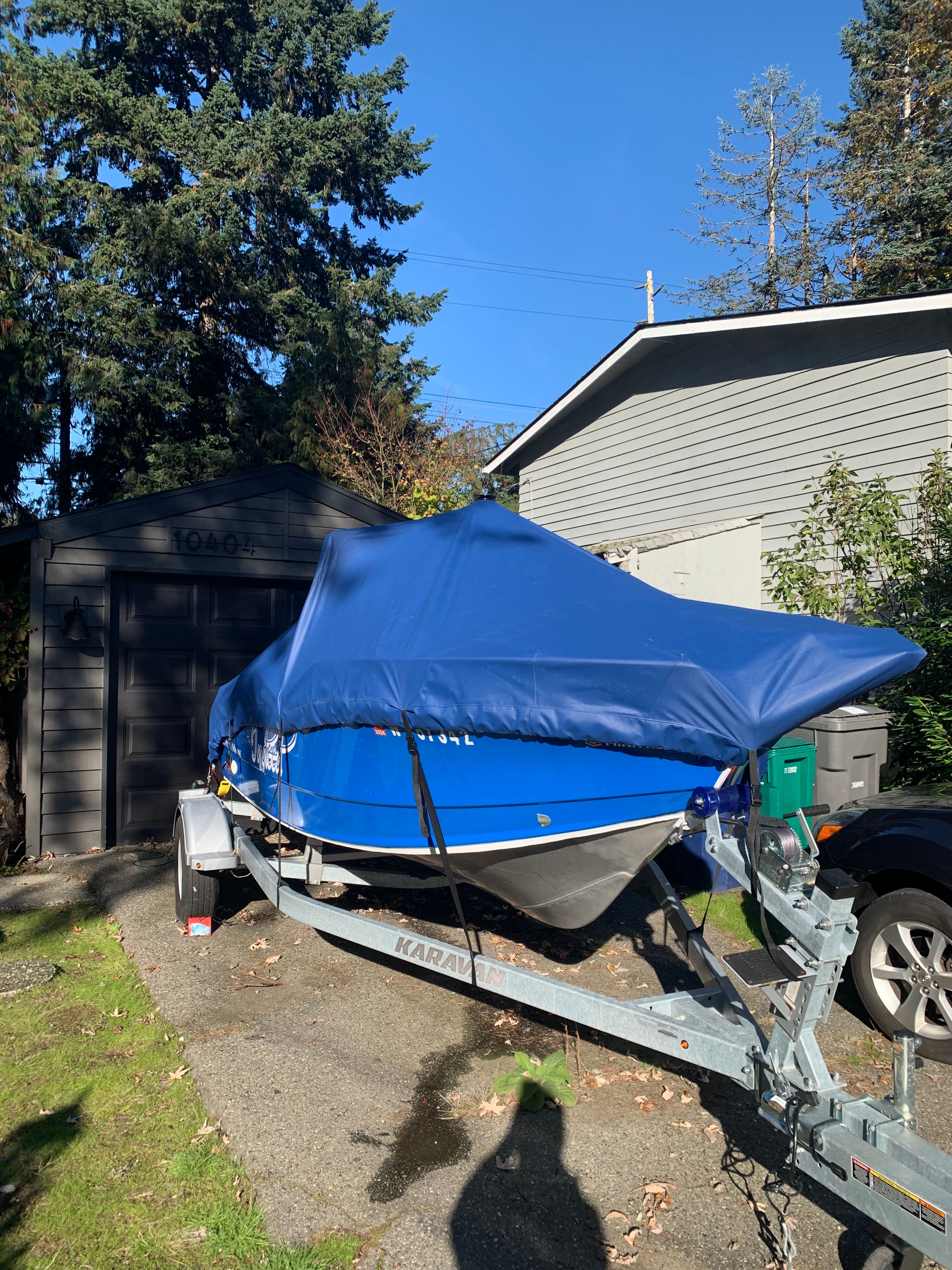 A blue boat on a trailer, covered with a tarp, is parked in a driveway.