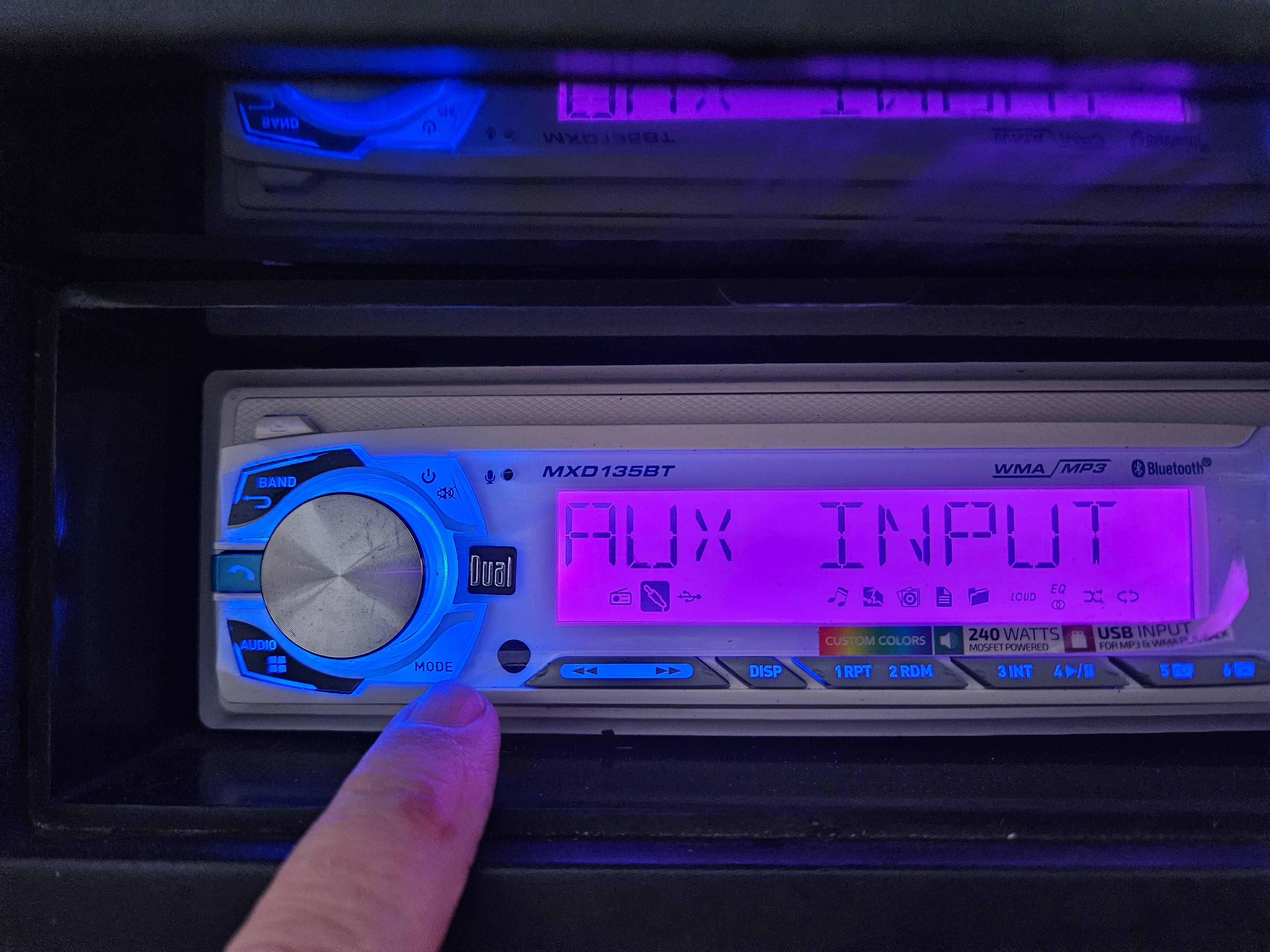 A finger points to a car stereo with a glowing display and AUX input.