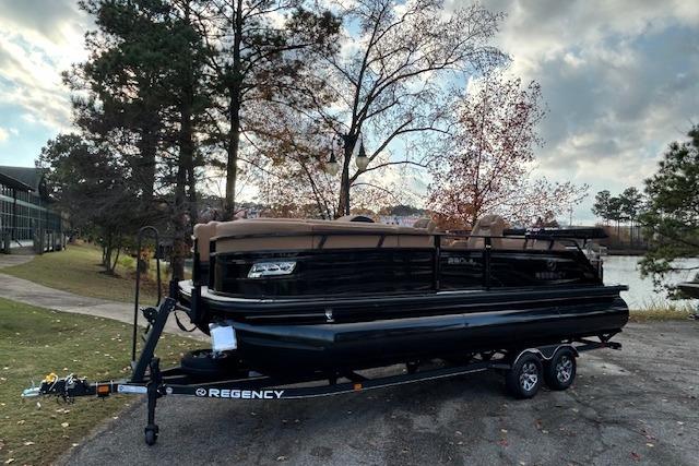 2025 Regency 230 LE3 Sport pontoon boat on trailer by lakeside, surrounded by trees.