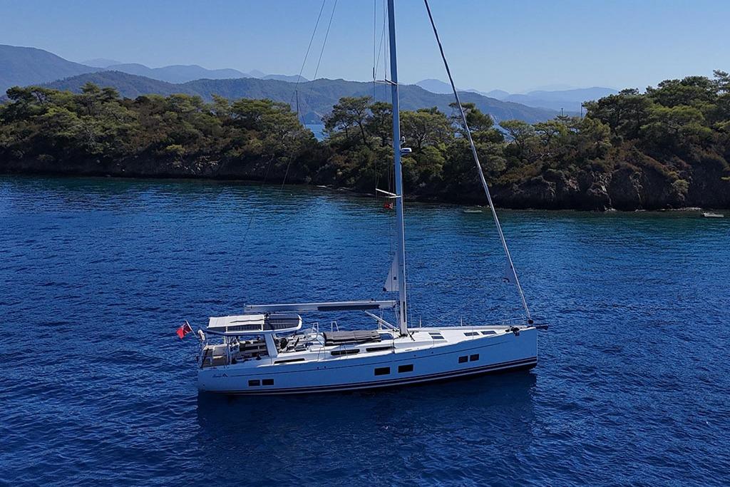 2024 Hanse 588 yacht sailing in clear blue waters near a lush, rocky coastline.