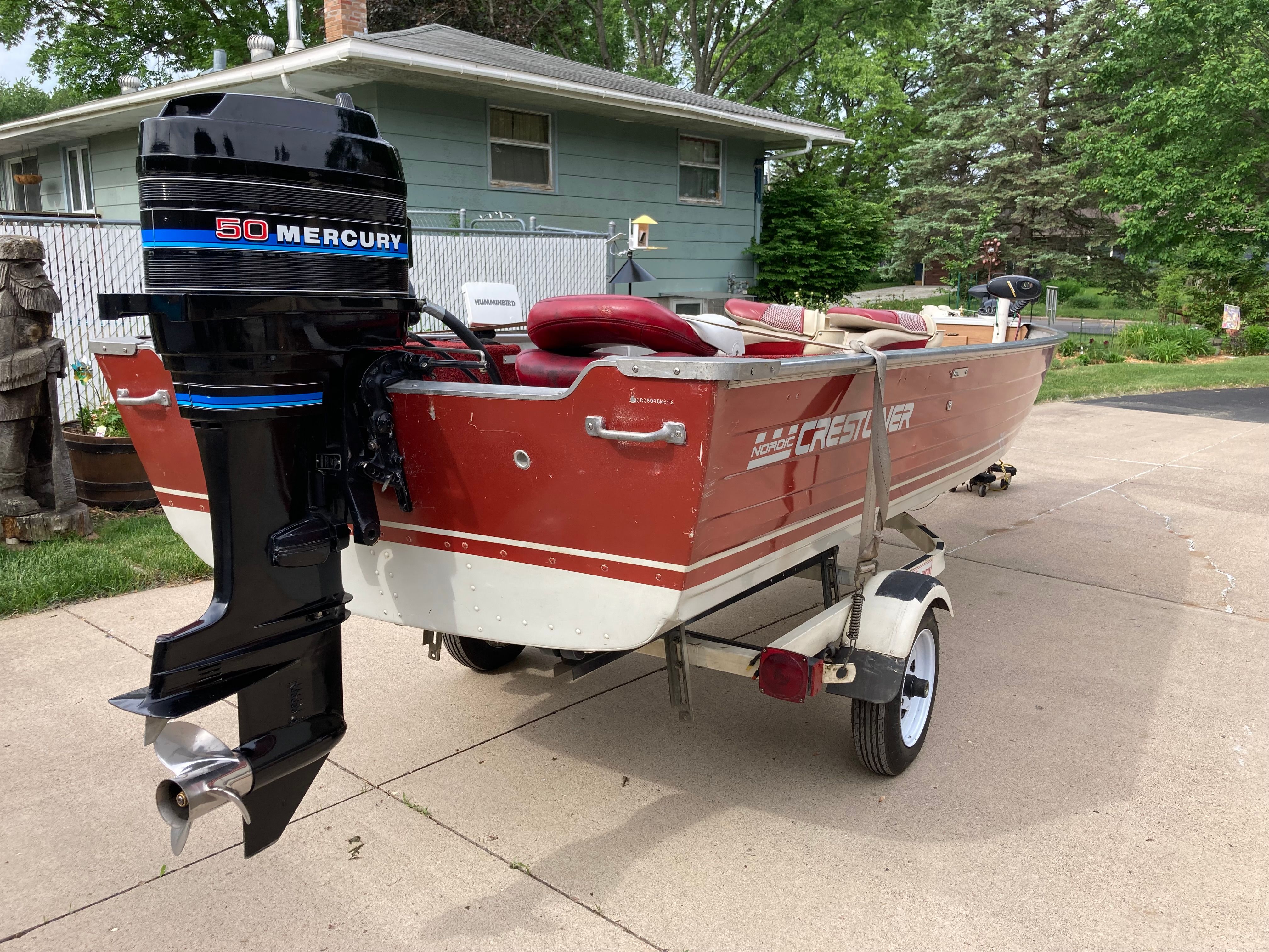 A red Crestliner boat with a Mercury engine on a trailer in a driveway.