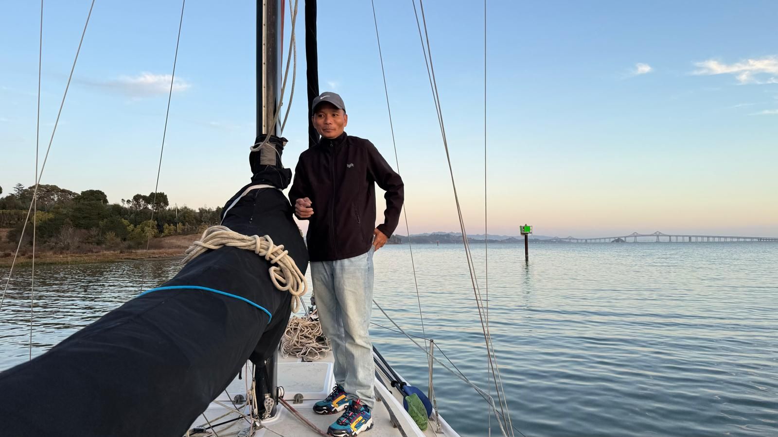 Person standing on a sailboat with calm water and a bridge in the background.