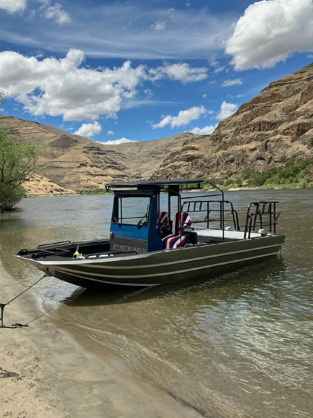 A Custom Weld 23' sled t-top boat is moored at a riverbank with a mountainous backdrop.
